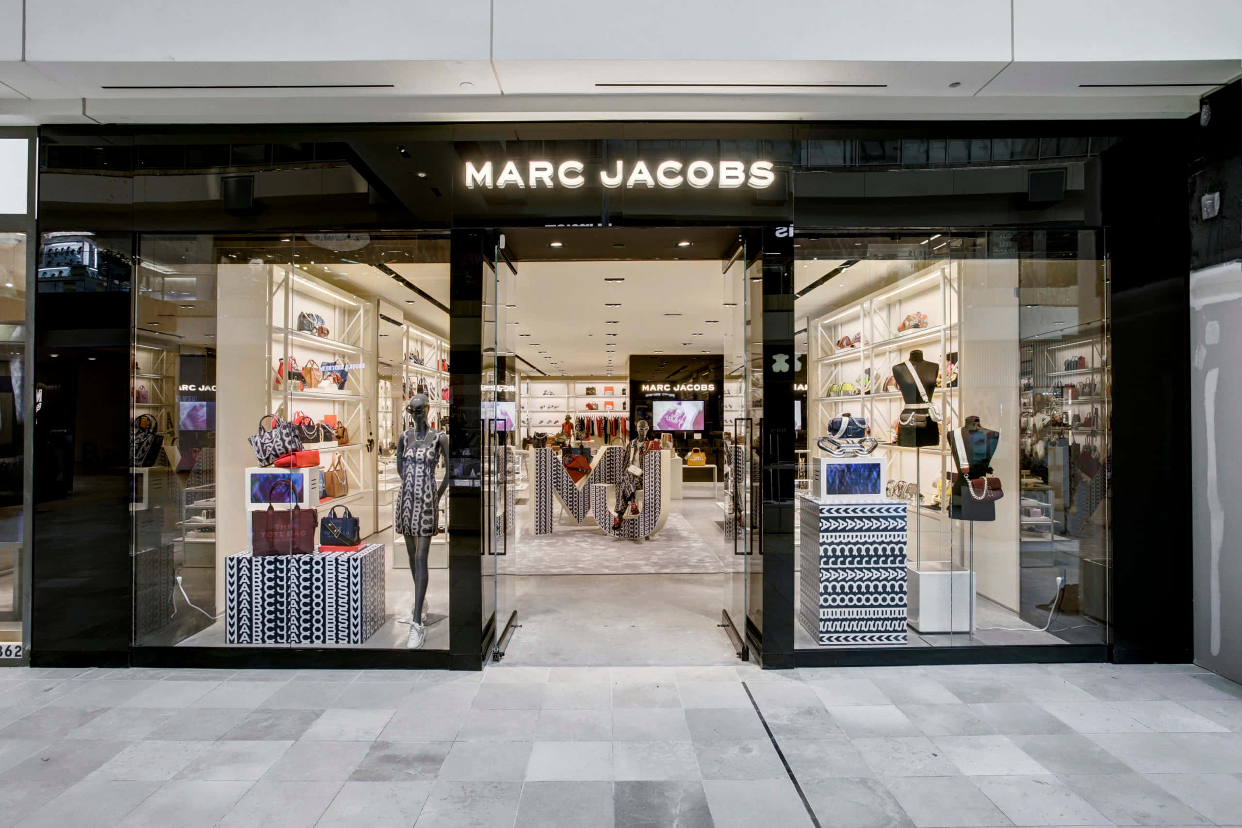 馬克·雅可布 桌布 壁紙 — 時尚的Marc Jacobs店鋪門面，配備玻璃門窗，展示人體模型和手提包，營造現代零售環境。