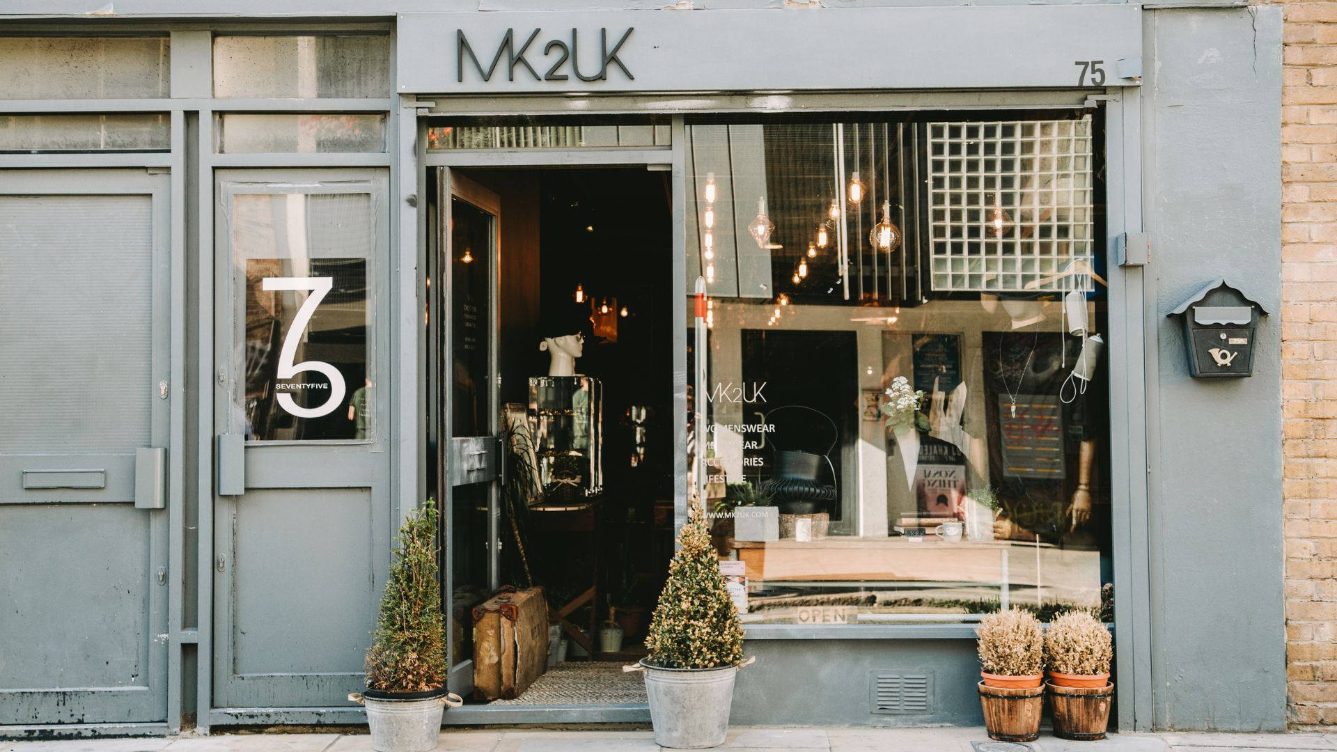 Store 桌布 壁紙 — 時尚的灰色店面，帶有「MK2UK」標誌和入口兩側的盆栽植物，顯露出溫暖的室內燈光和復古裝飾。