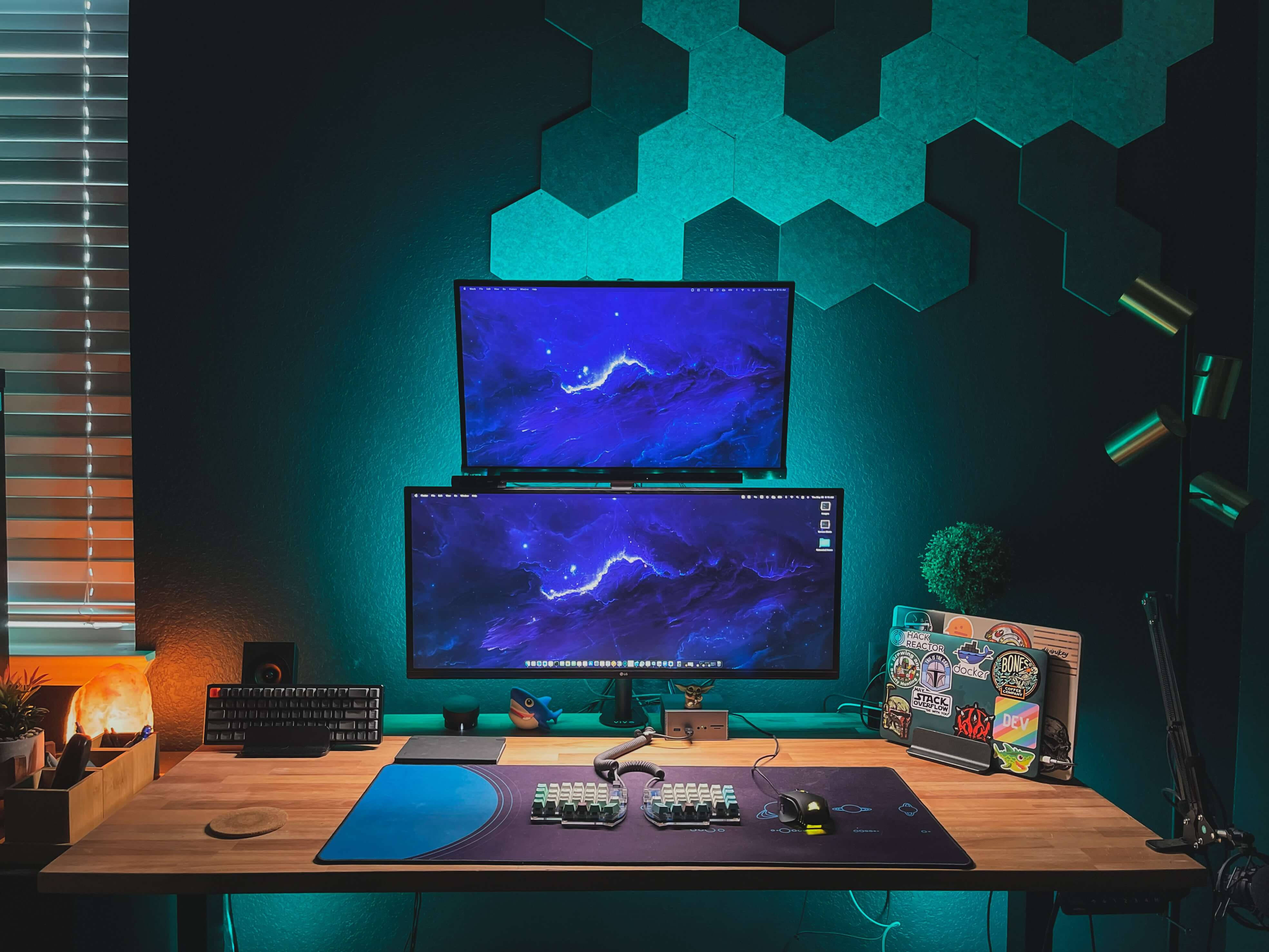 Desk 桌布 壁紙 — 時尚的遊戲設置，配備雙顯示器，在青色環境照明和六邊形牆板下顯示宇宙壁紙。