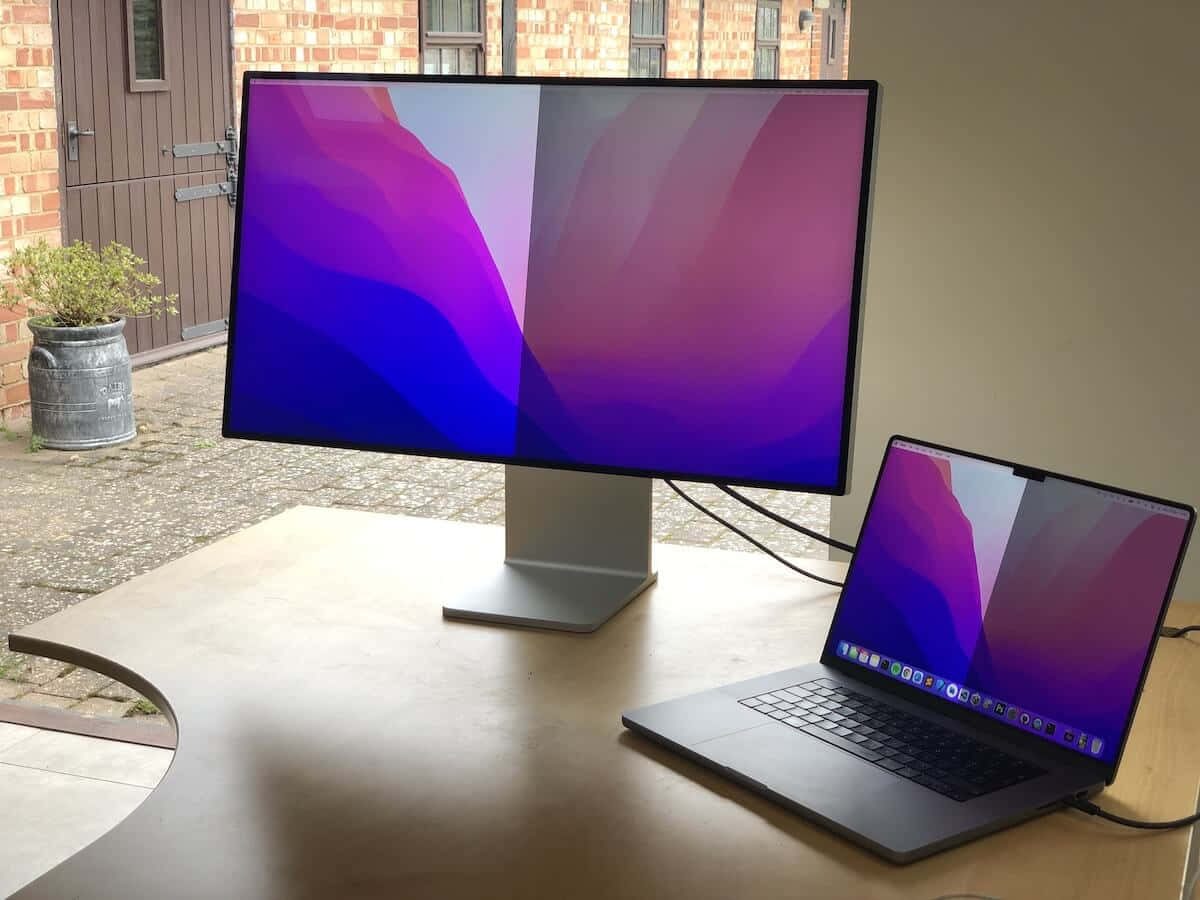 MacBook 動態圖片 桌布 壁紙 — 時尚的銀色顯示器和筆記型電腦在磚砌建築附近的木桌上展示充滿活力的紫色、藍色和粉紅色抽象桌布。