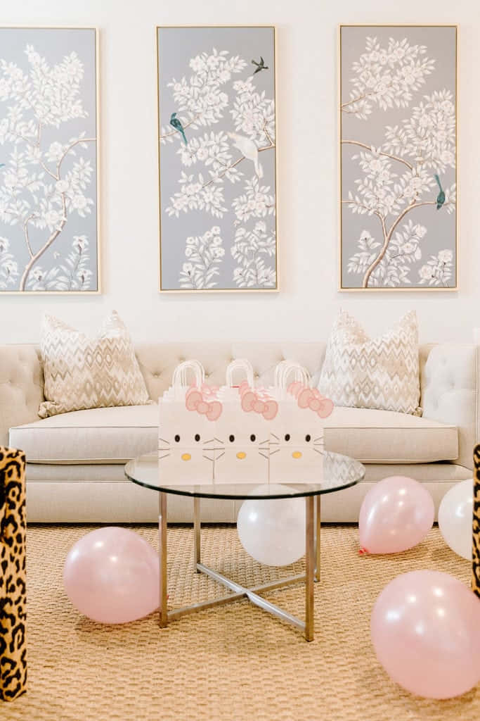 Hello Kitty Party 桌布 壁紙 — 時尚育嬰室，配有米色沙發、圖案抱枕，上方掛有三幅裱框植物畫，周圍環繞著粉紅色和白色氣球以及Hello Kitty禮物袋。