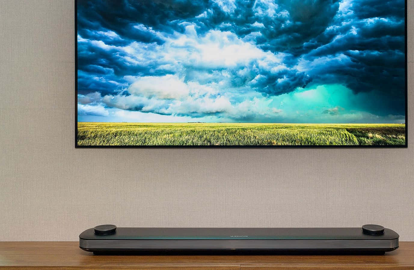 tv 桌布 壁紙 — 時尚黑色 soundbar 放置在木架上，下方的大型電視顯示金色田野上空戲劇性的暴風雨天空。