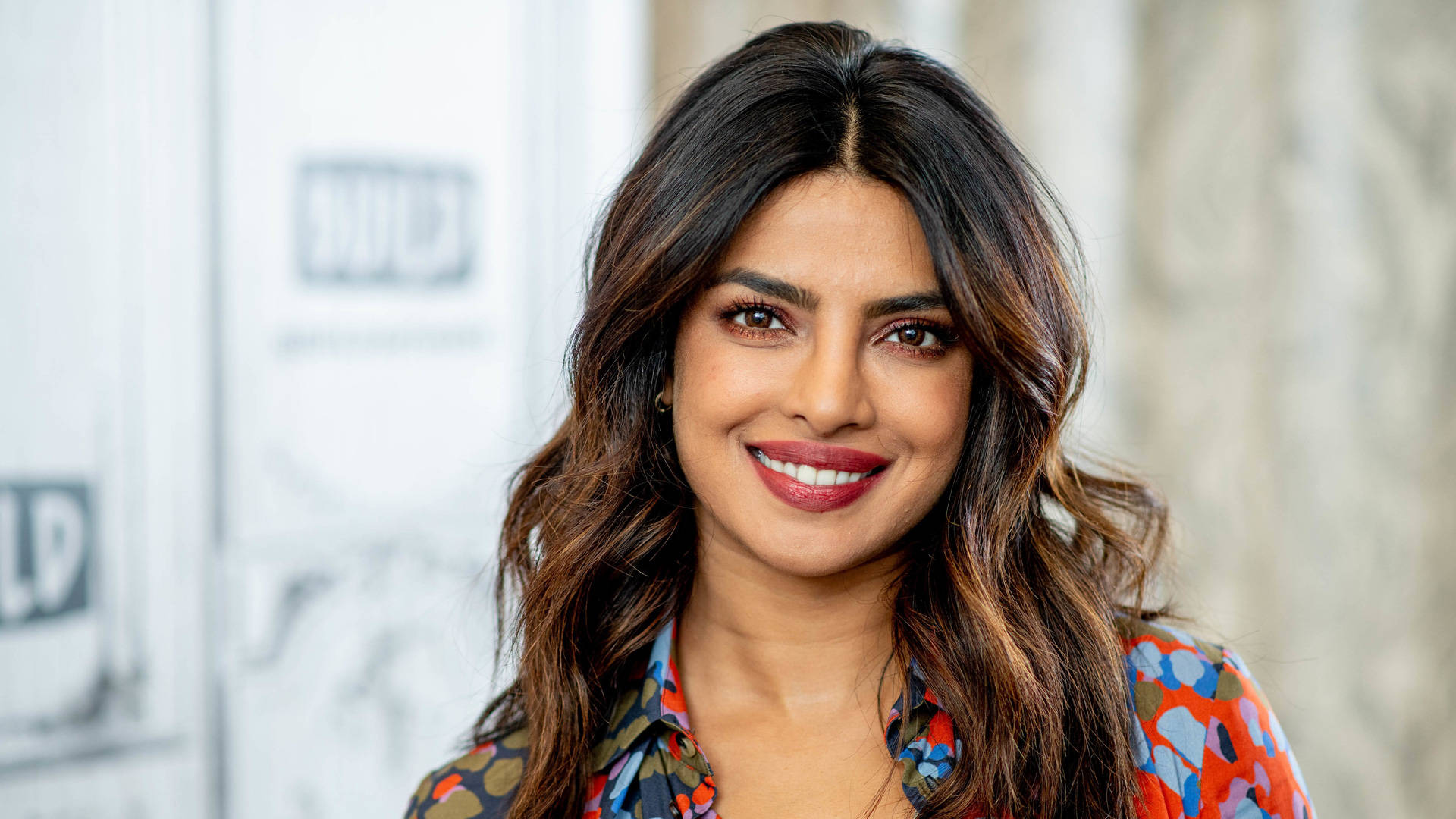 Priyanka 桌布 壁紙 — 普里揚卡·喬普拉在模糊的活動背景下，穿著色彩繽紛的圖案襯衫，溫暖地微笑。