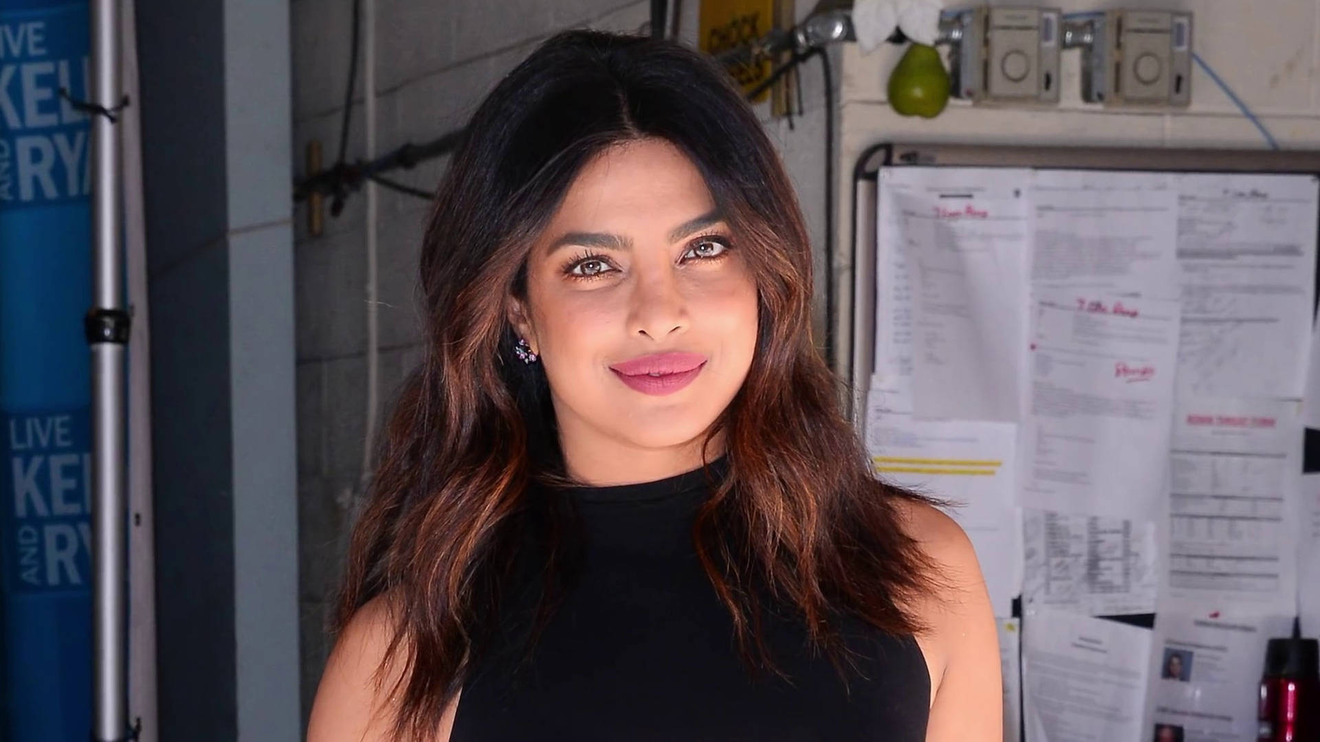 Priyanka 桌布 壁紙 — 普里揚卡·喬普拉身穿黑色上衣，在雜亂的後台環境中溫暖微笑，周圍有紙張和設備。