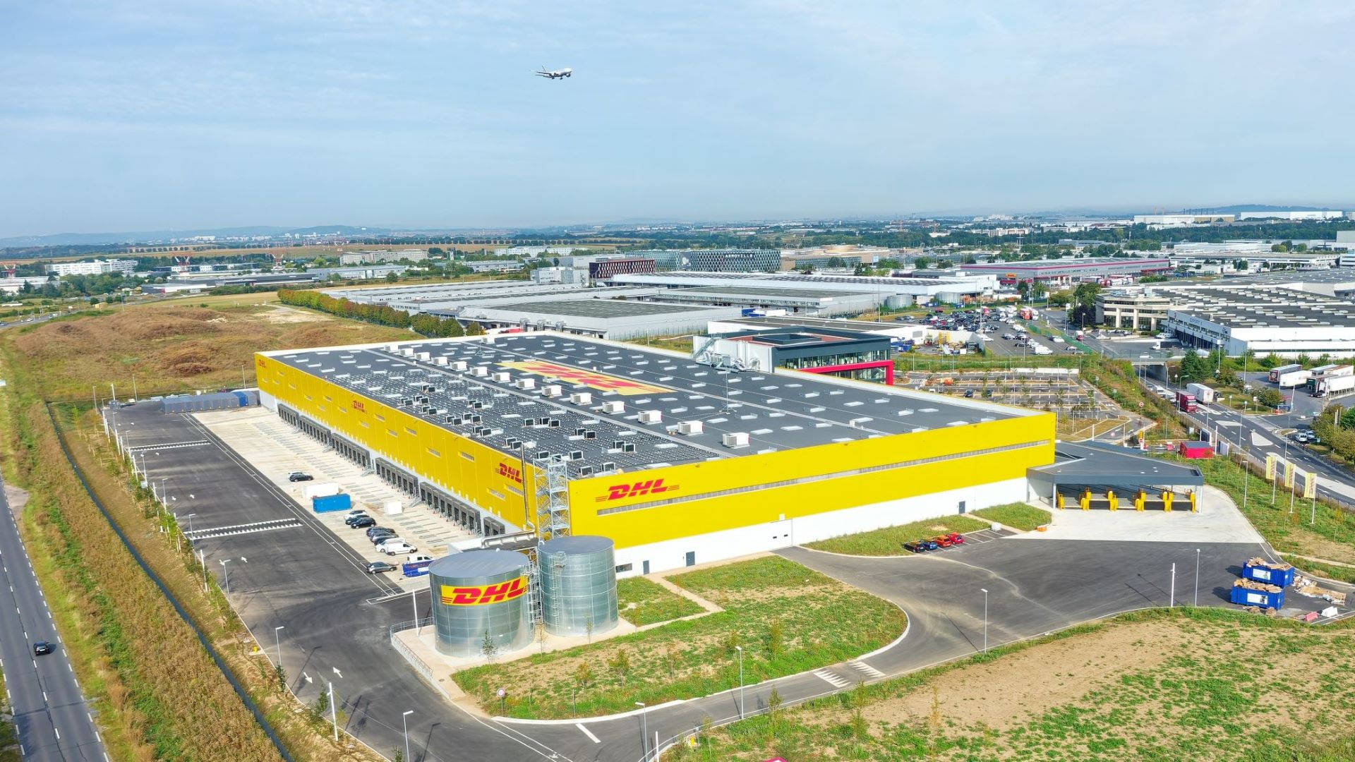 Dhl 桌布 壁紙 — 晴朗天空下，一個巨大的黃色DHL物流倉庫，灰色屋頂及周邊道路的鳥瞰圖。
