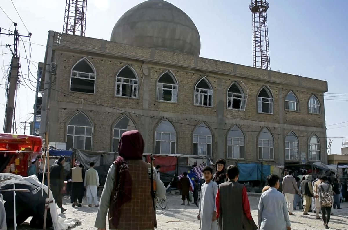 Taliban 桌布 壁紙 — 晴朗天空下，一座受損清真寺前熱鬧的街景，清真寺有大型圓頂和拱形窗戶。