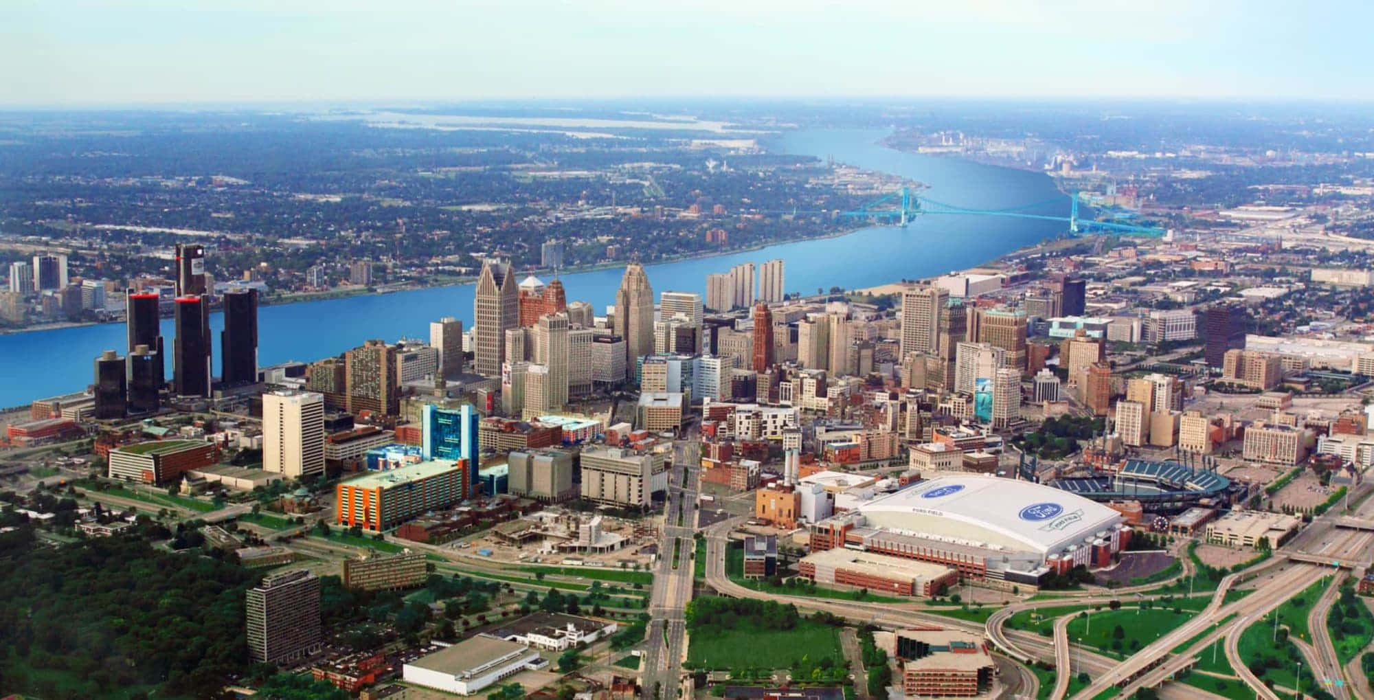 Michigan 桌布 壁紙 — 晴朗天空下，寬闊河流與橋樑穿過密集城市建築的廣闊城市景觀鳥瞰圖。