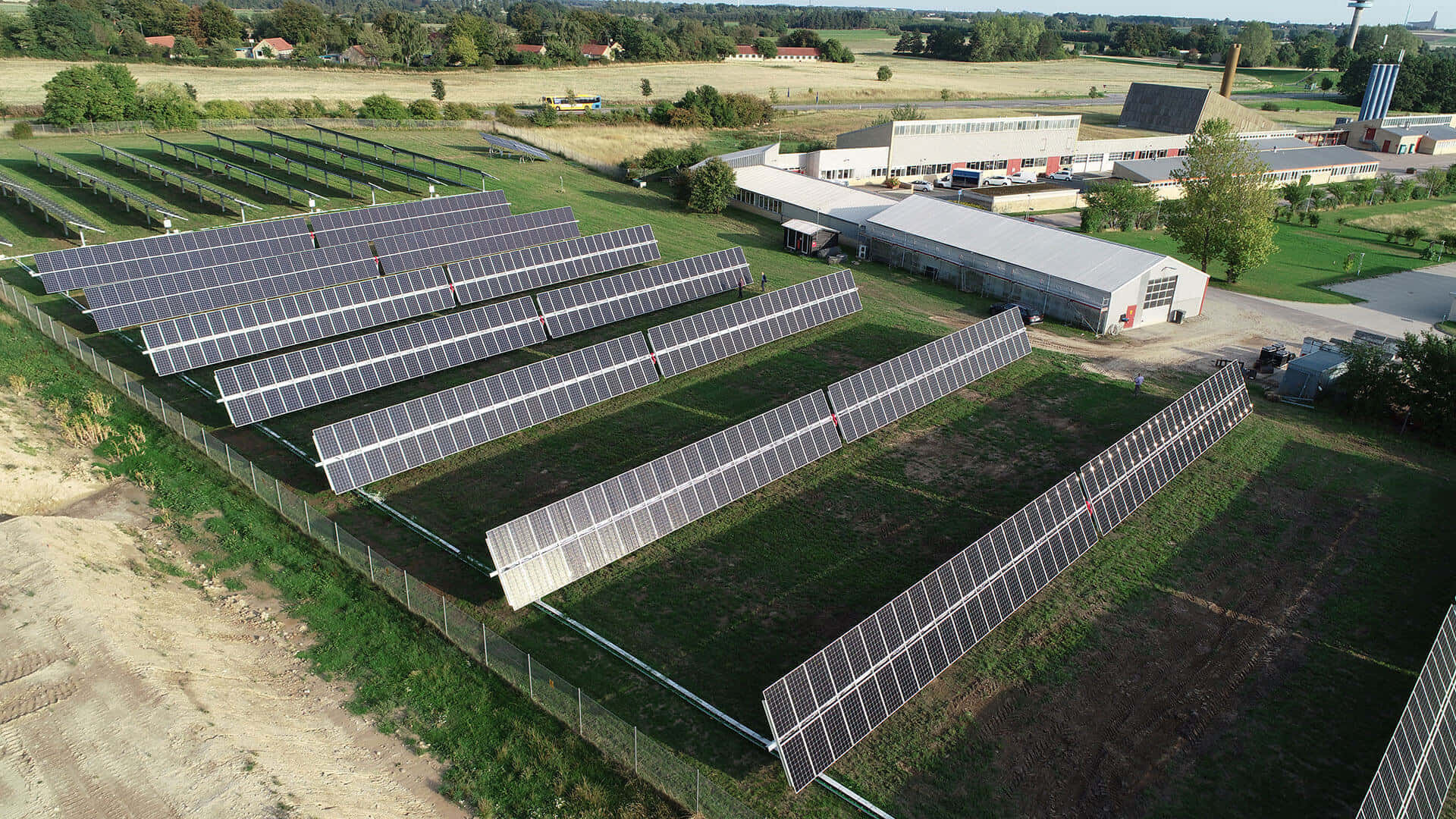 Denmark 桌布 壁紙 — 晴朗天空下，工業建築物旁邊的綠色田野上整齊排列的大型太陽能電池板陣列的鳥瞰圖。