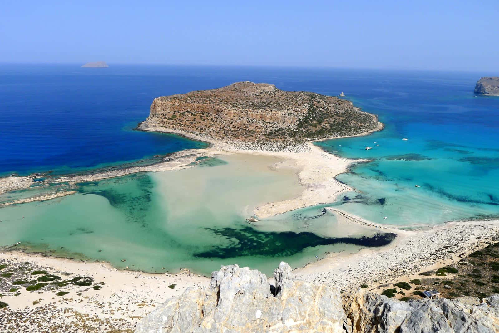 Greek Beach 桌布 壁紙 — 晴朗天空下，深藍色海洋環繞著一個擁有碧綠潟湖和白色沙灘的迷人島嶼的鳥瞰圖。