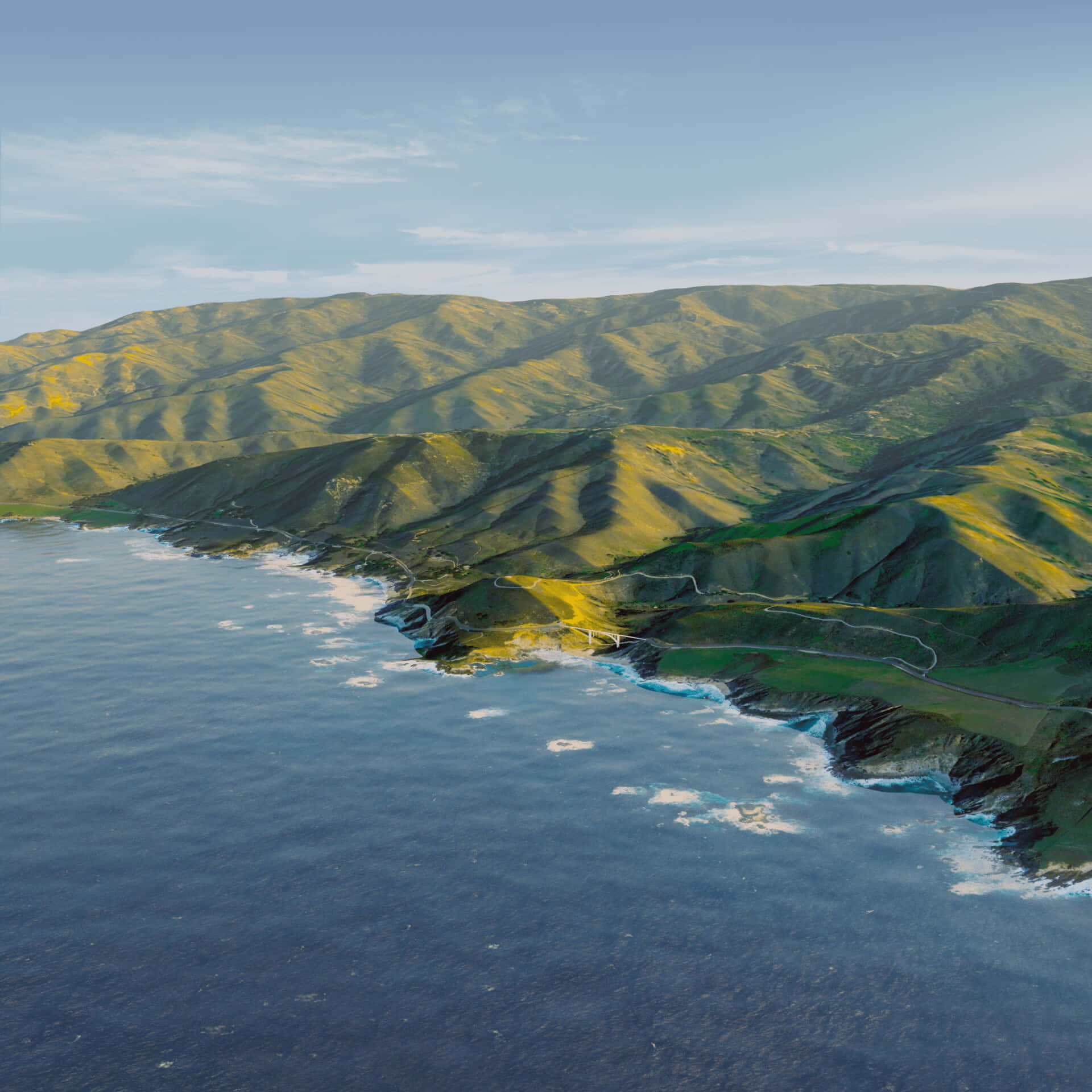 macOS Big Sur 動態桌布 桌布 壁紙 — 晴朗天空下，蜿蜒海岸公路擁抱著翠綠山丘，與深藍海洋交匯的鳥瞰圖。
