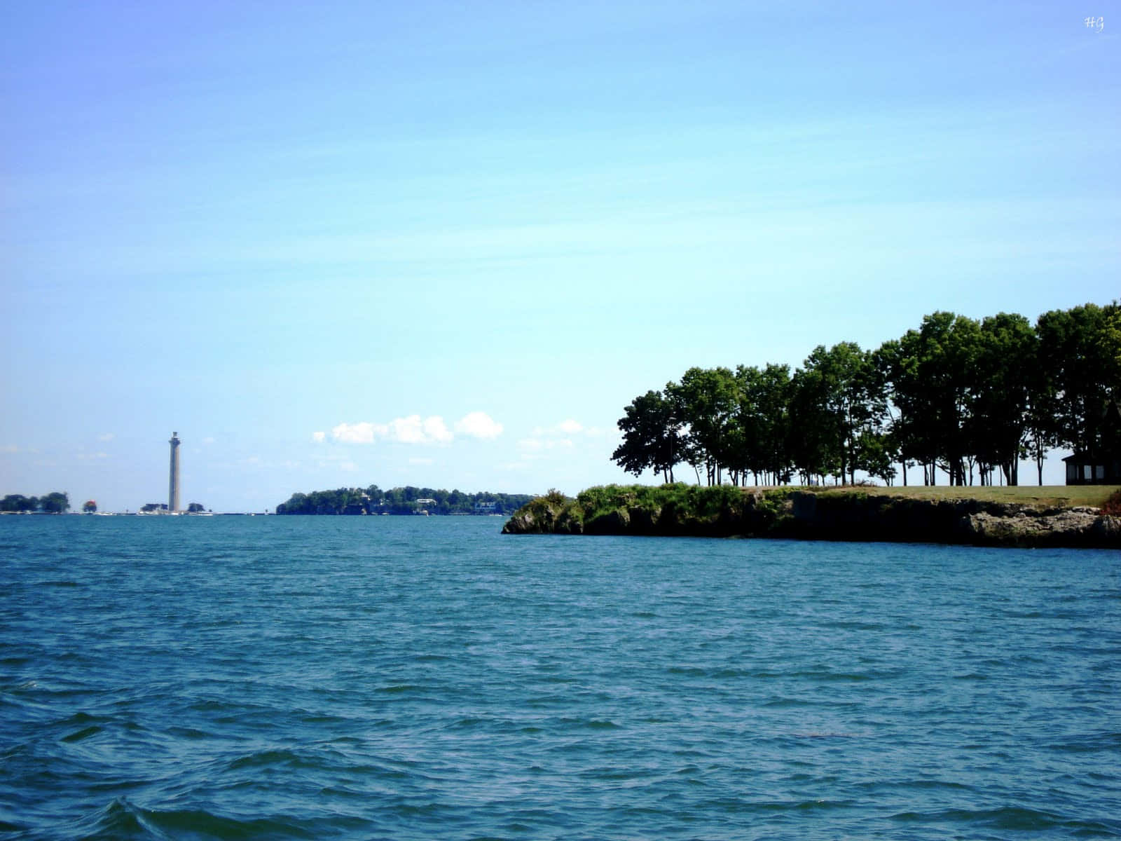Lake Erie 桌布 壁紙 — 晴朗天空下，遠處有燈塔和樹木成蔭的岸邊的寧靜藍色水景。