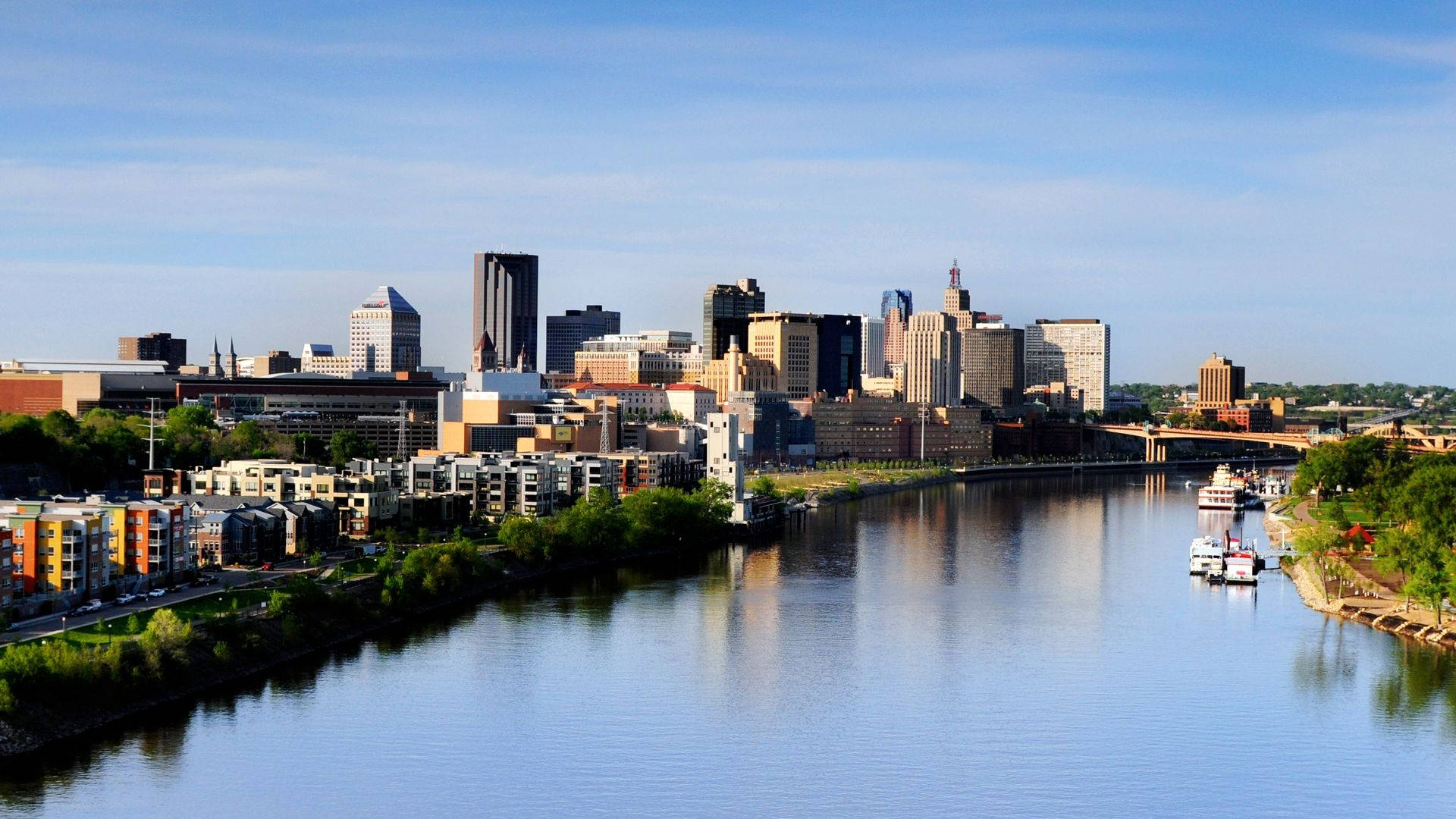 Minnesota 桌布 壁紙 — 晴朗日子裡，現代城市天際線倒映在平靜的河面上，河岸有船隻和綠樹。