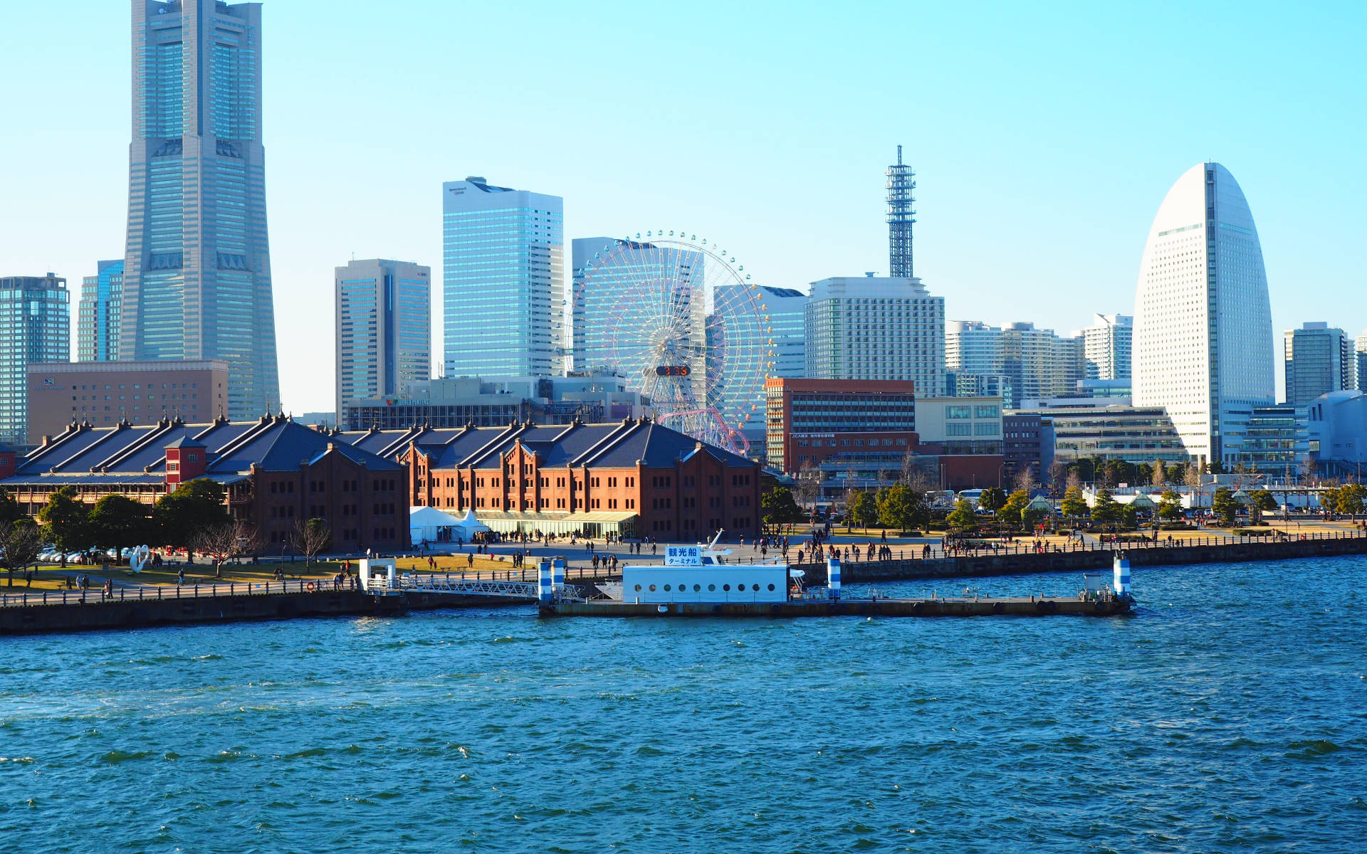 橫濱 桌布 壁紙 — 晴朗日子裡現代城市天際線的景色，高聳的玻璃塔和大型摩天輪背後是歷史悠久的紅磚倉庫，沿著藍色海濱。