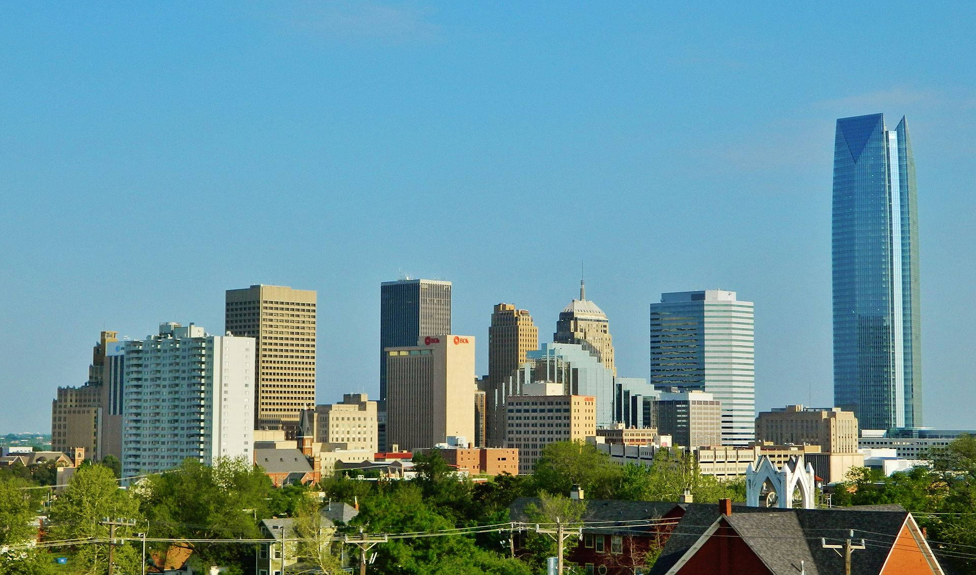 Oklahoma 桌布 壁紙 — 晴朗明媚的一天，展示了現代城市天際線，玻璃摩天大樓聳立在綠樹和紅頂房屋之上。