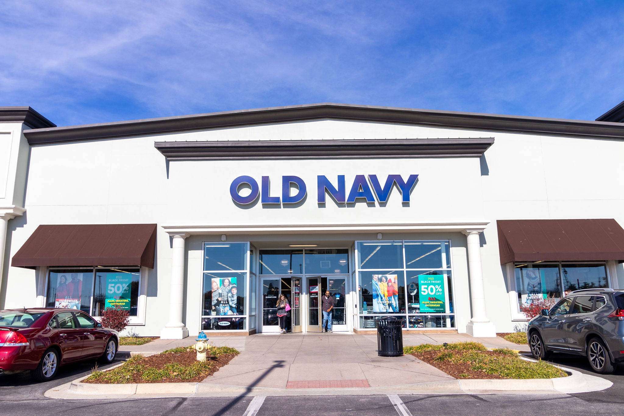 old 桌布 壁紙 — 晴朗的一天，Old Navy 商店內有人從玻璃門進入，外面停著汽車，天空清澈湛藍。