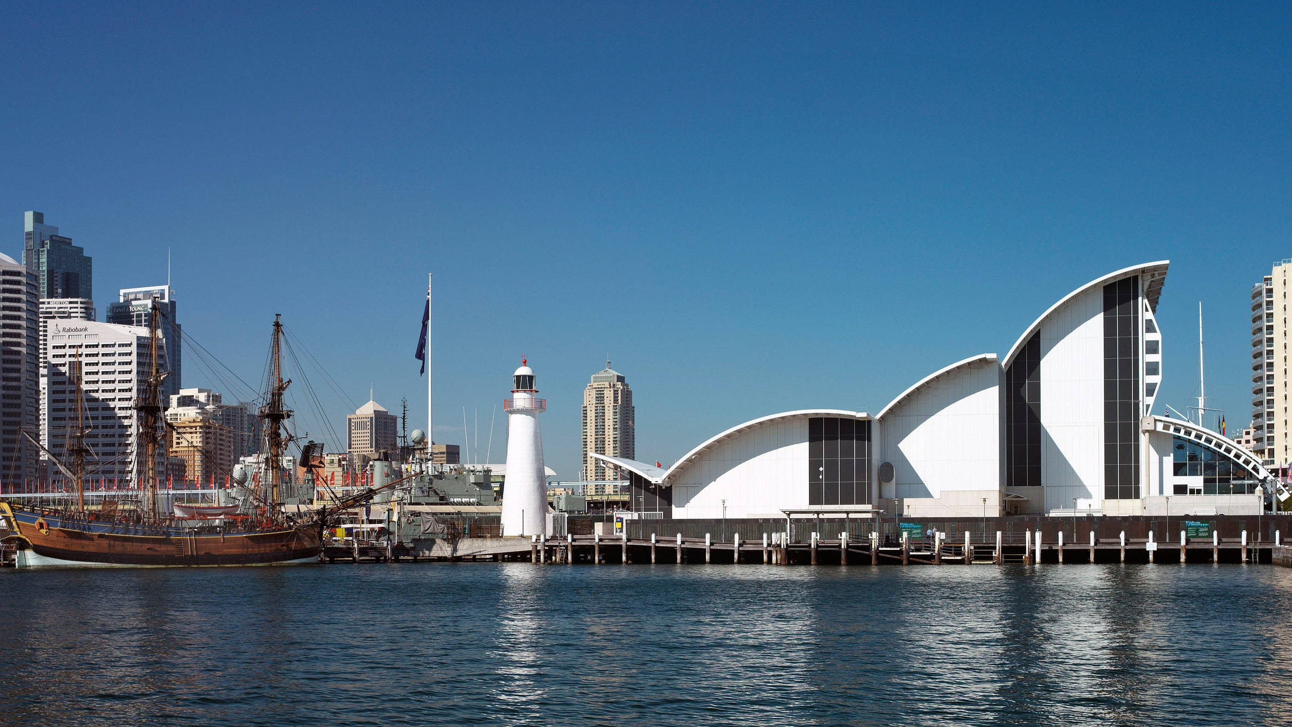 Sydney 桌布 壁紙 — 晴朗的一天，繁忙的港口中，一艘歷史悠久的船隻停泊在現代白色曲線建築和燈塔旁邊，天空清澈湛藍。