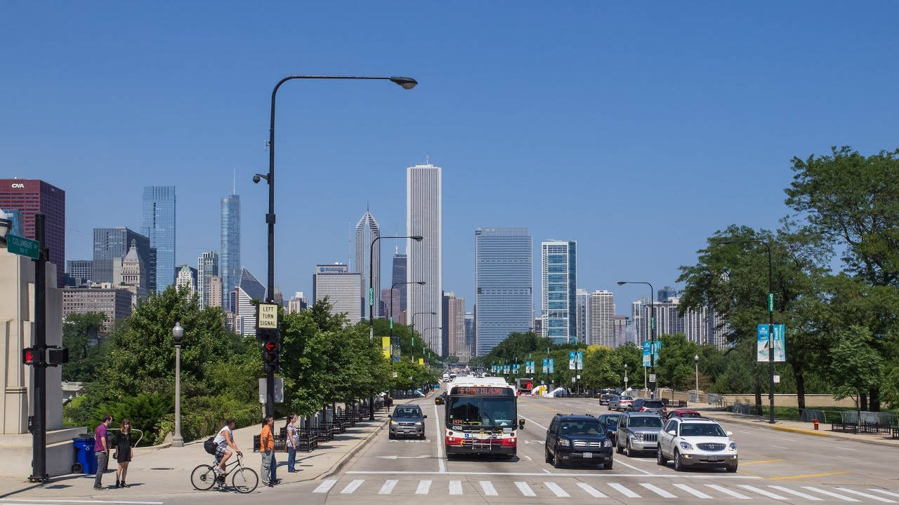 City Street 桌布 壁紙 — 晴朗的城市街道，汽車和騎自行車的人在高聳的城市天際線前，背景是清澈的藍天。