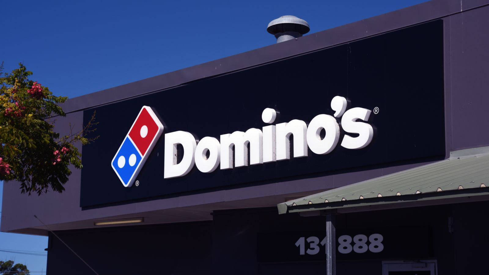 Dominos Pizza 桌布 壁紙 — 晴朗的藍天襯托著 Domino's Pizza 店面，白色粗體字和紅藍標誌在清晰日光下格外醒目。