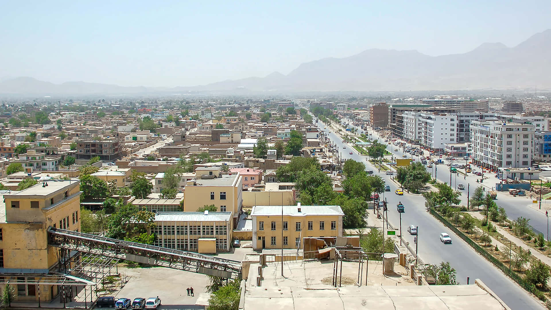 Afghanistan 桌布 壁紙 — 晴朗藍天下，一個擁有蜿蜒街道、汽車和遠處山脈的廣闊城市的鳥瞰圖。
