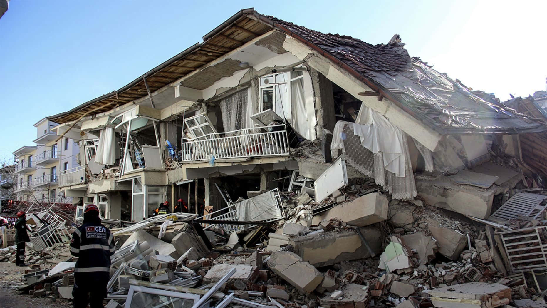Earthquake 桌布 壁紙 — 晴朗藍天下，倒塌建築物散落破碎混凝土與瓦礫，救援人員正在評估損毀情況。