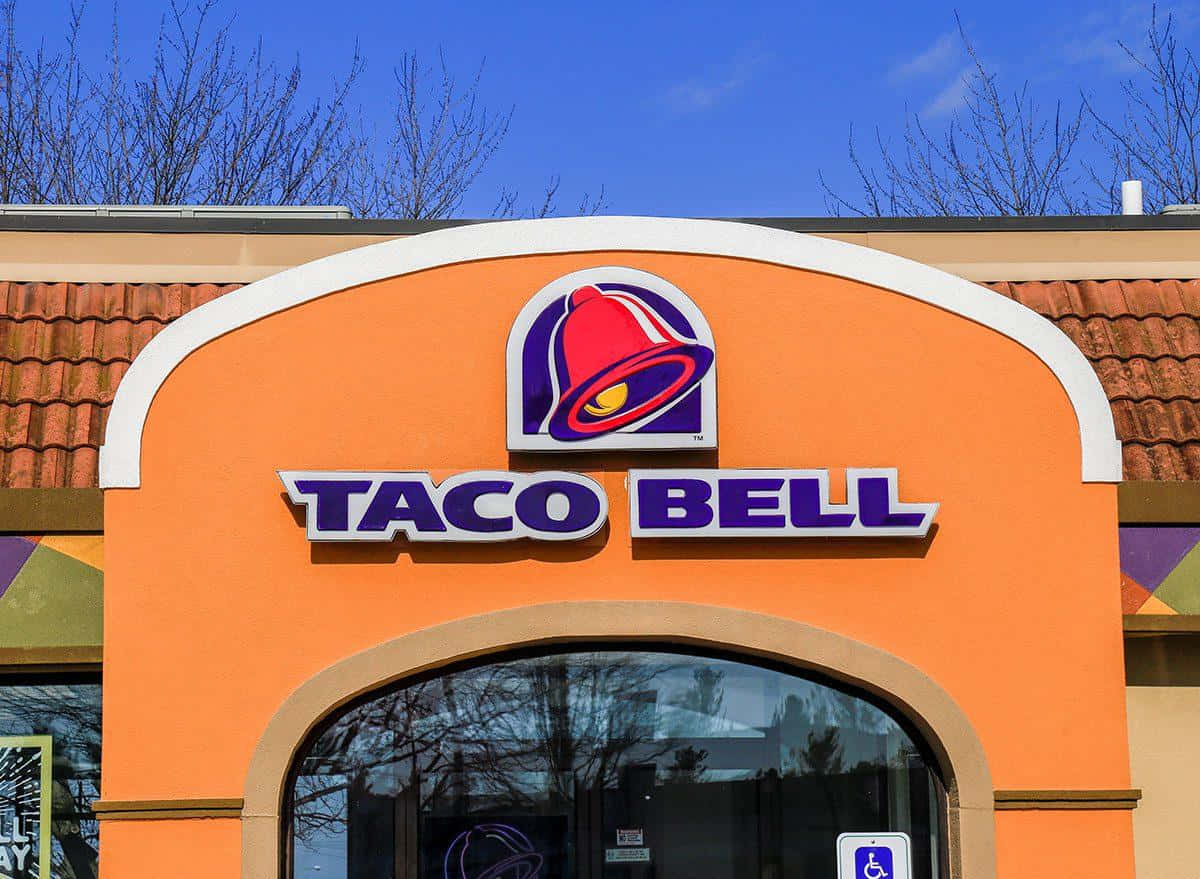 塔可鐘 桌布 壁紙 — 晴朗藍天下，帶有裸露樹枝的鮮橙色Taco Bell餐廳外牆。