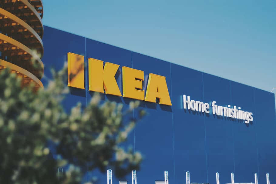 Ikea 桌布 壁紙 — 晴朗藍天下，明亮的藍色IKEA商店外牆搭配醒目的黃色標誌，前景有模糊的綠色樹葉作為框景。