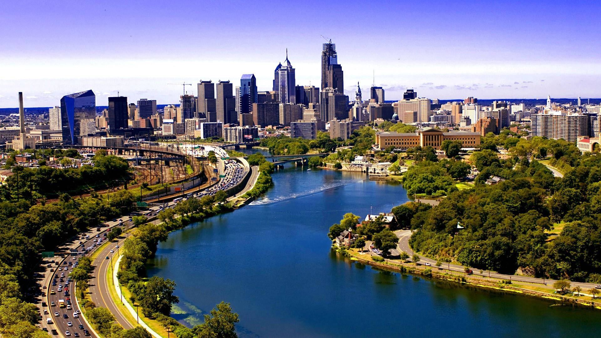 Philadelphia 桌布 壁紙 — 晴朗藍天下，蜿蜒河流兩旁綠意盎然的公園和繁忙高速公路交織的活力城市天際線鳥瞰圖。