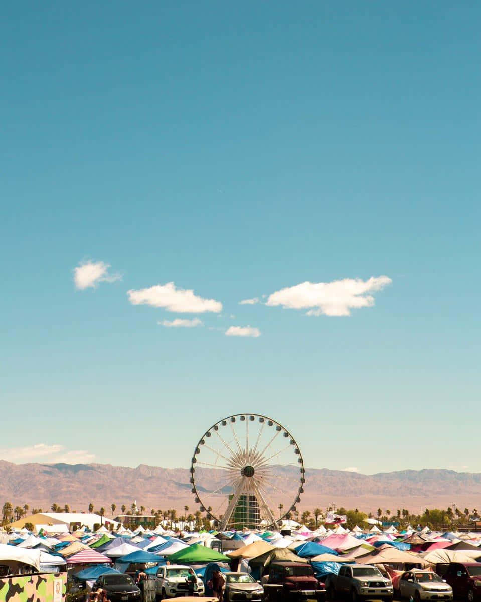 Coachella 桌布 壁紙 — 晴朗藍天下充滿活力的沙漠節日，巨大的摩天輪聳立在色彩繽紛的帳篷和停泊的汽車之上。
