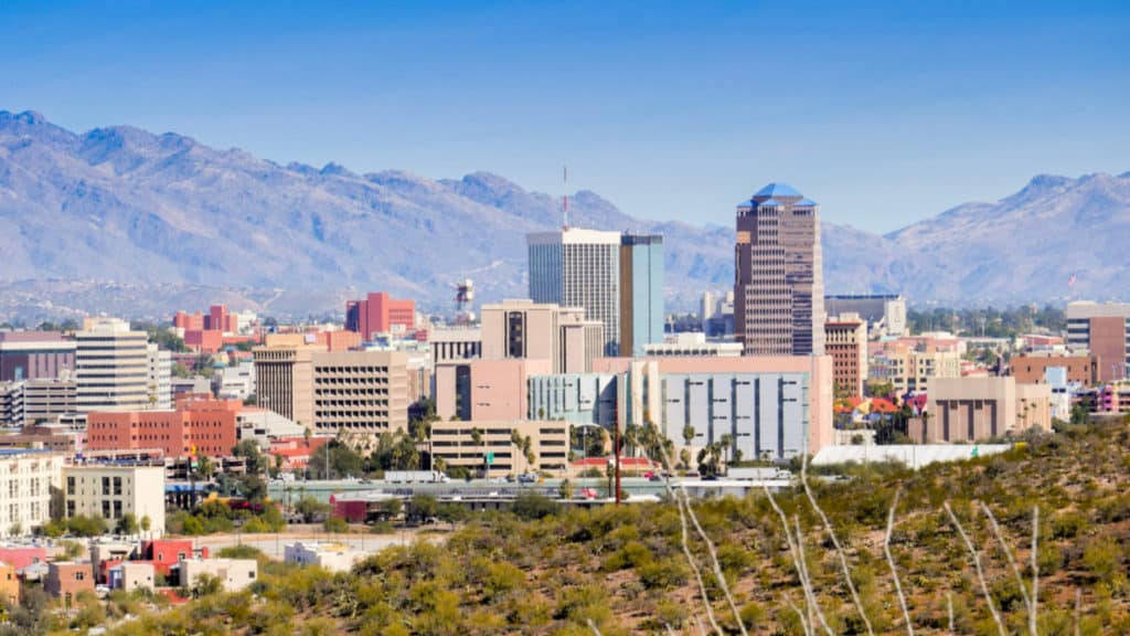 Arizona 桌布 壁紙 — 晴朗陽光下的城市景觀，現代建築依偎在遠處崎嶇的山巒前，背景是清澈的藍天。