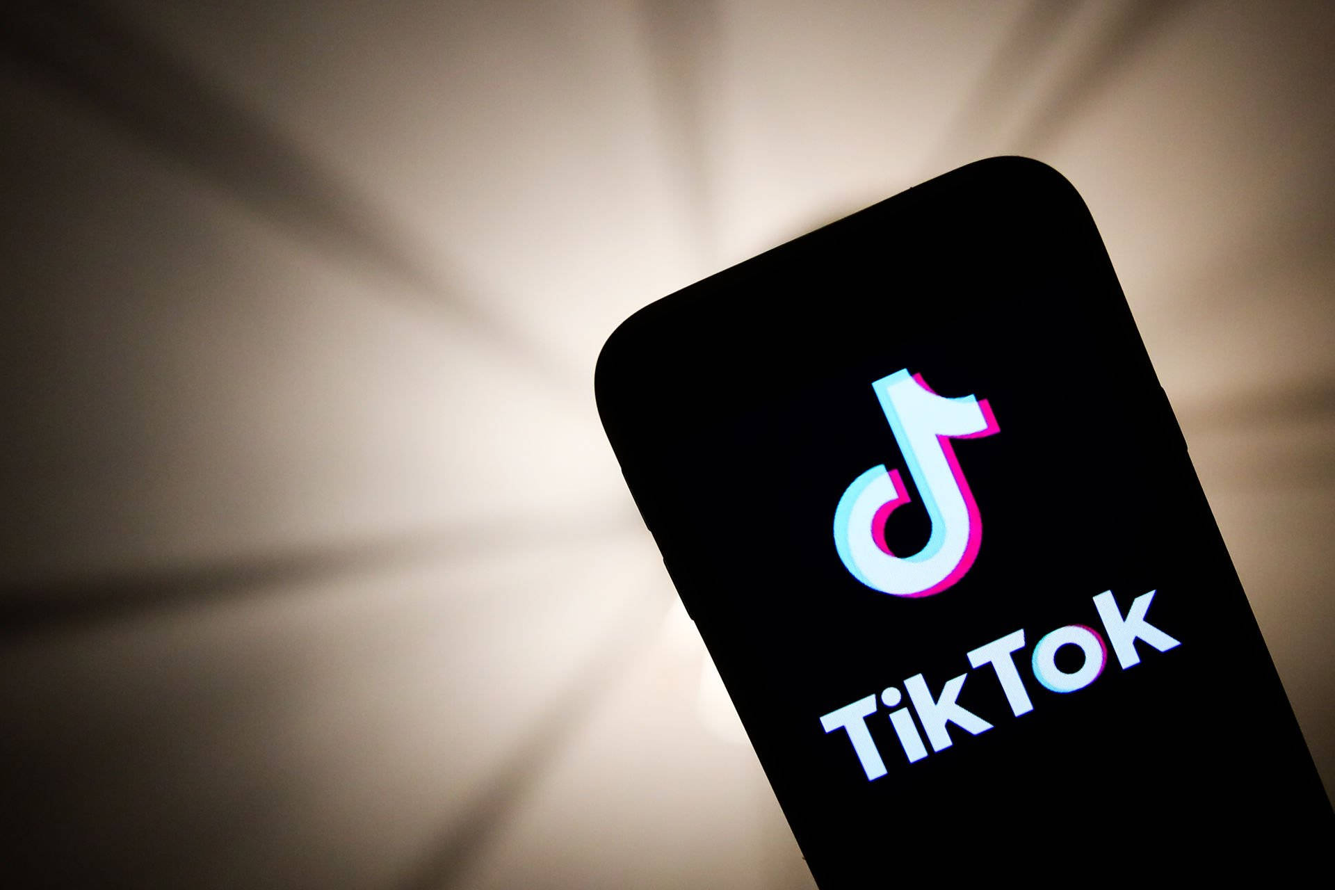 TikTok 動圖 桌布 壁紙 — 智慧型手機在柔和光線和放射陰影的背景上顯示發光的TikTok標誌。