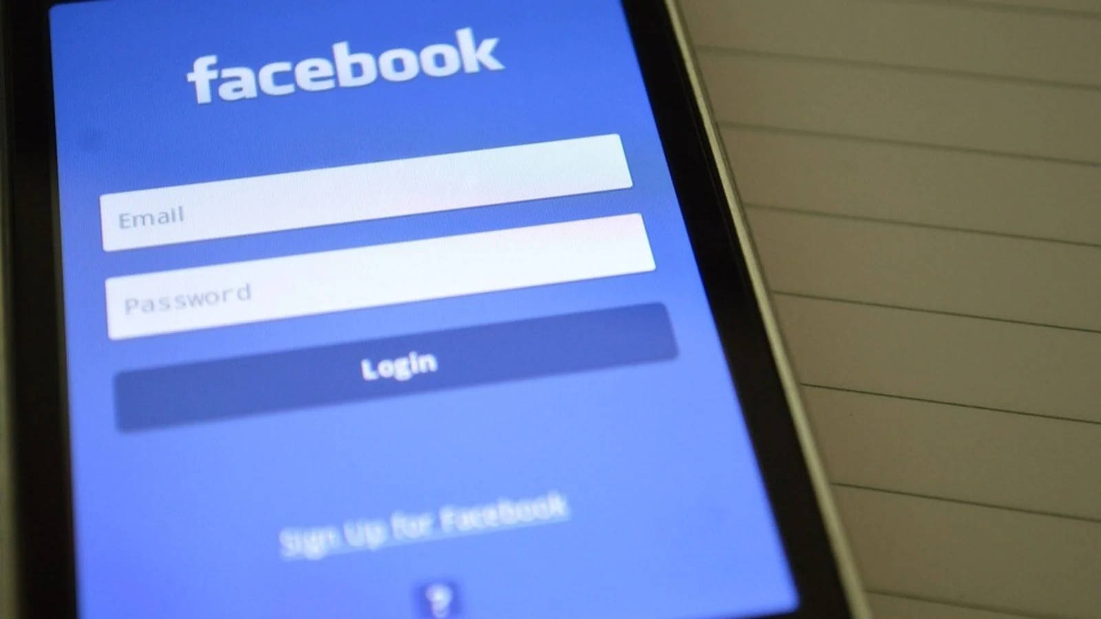 Facebook Login 桌布 壁紙 — 智慧型手機特寫，顯示Facebook登入畫面，包含電子郵件和密碼欄位，背景為橫線筆記本。