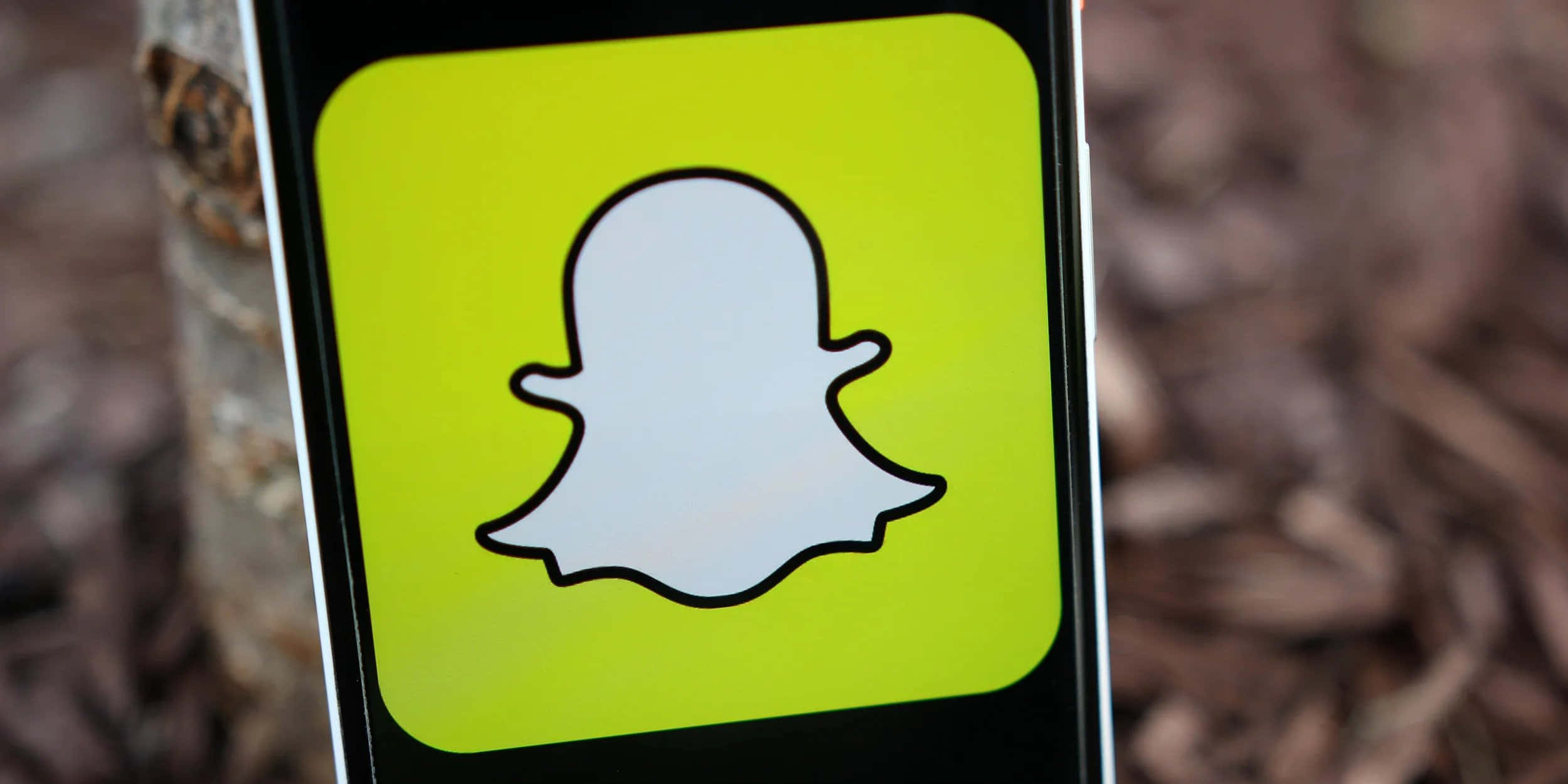 Snapchat 桌布 壁紙 — 智慧型手機特寫，顯示Snapchat鬼魂標誌，背景為模糊的自然景色。