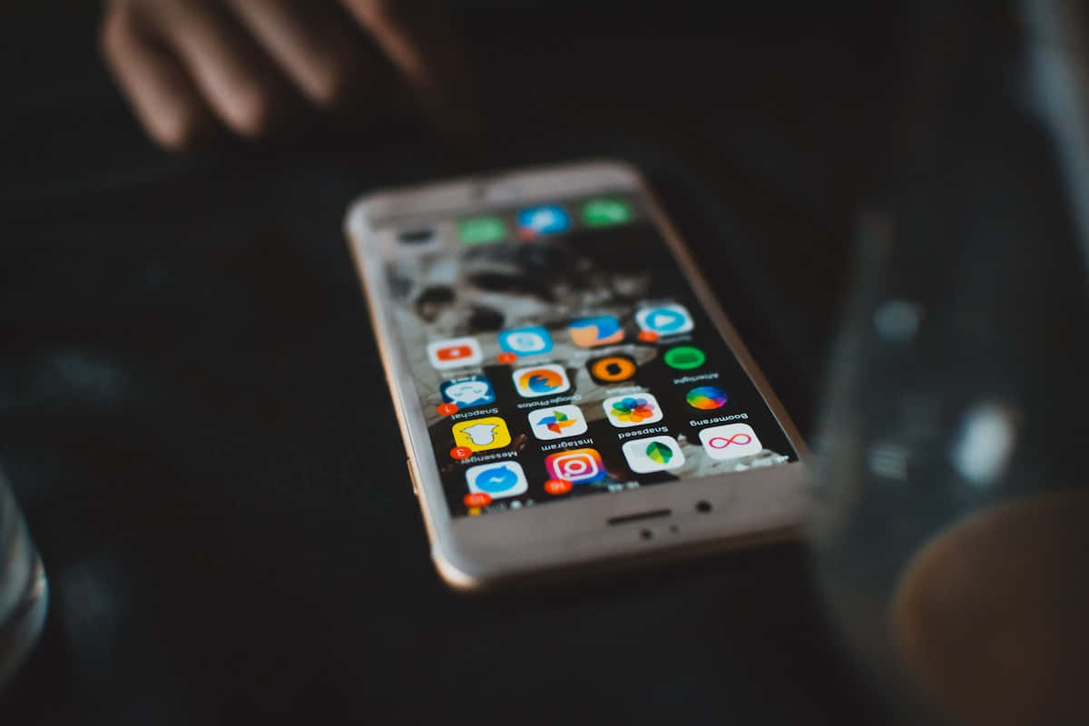 iPhone 桌布 壁紙 — 智慧型手機的特寫，在深色桌子上顯示色彩繽紛的應用程式圖示，背景有模糊的手和玻璃器皿。