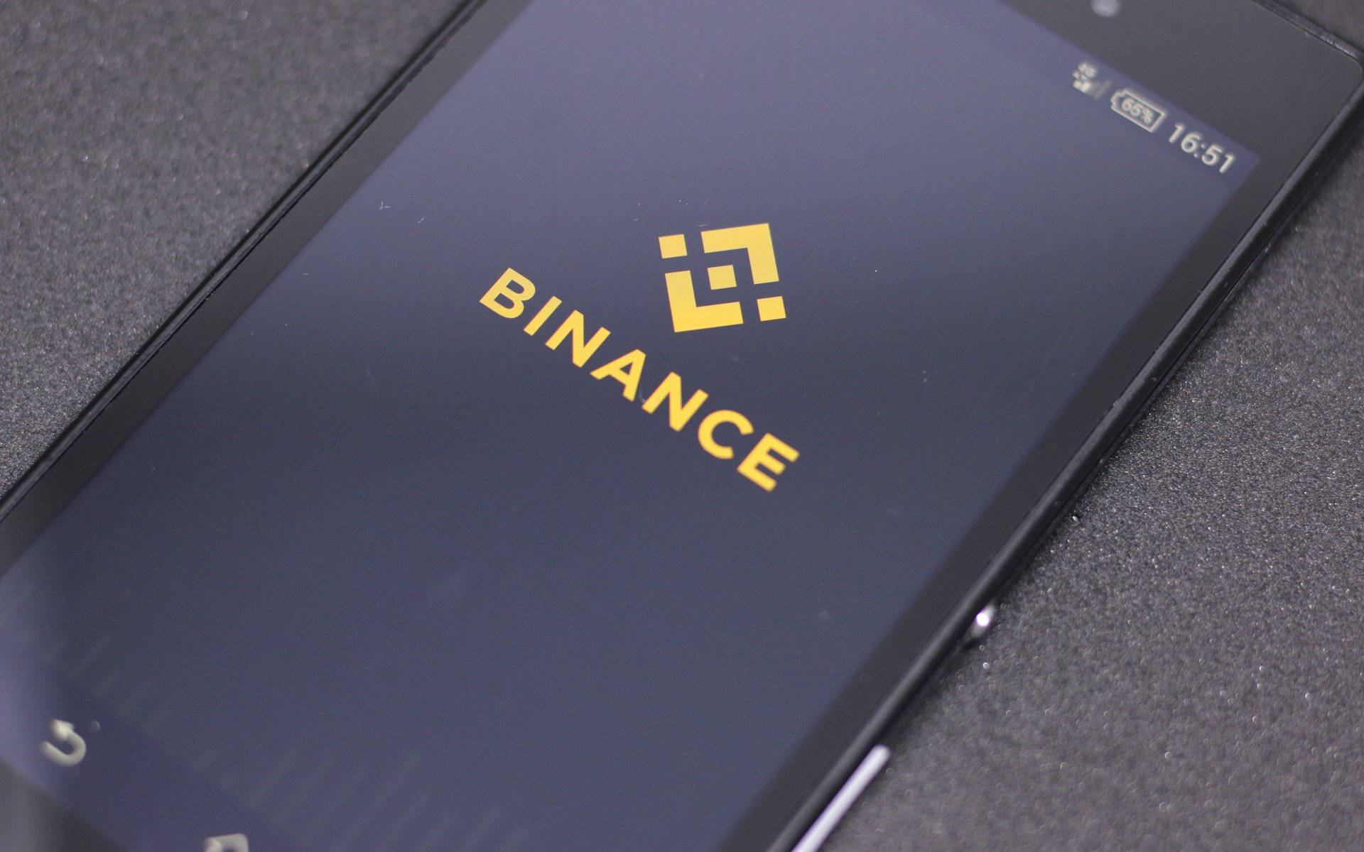 Binance 桌布 壁紙 — 智慧型手機的特寫照片，在深色螢幕上顯示金色的 Binance 標誌，放置在紋理灰色表面上。