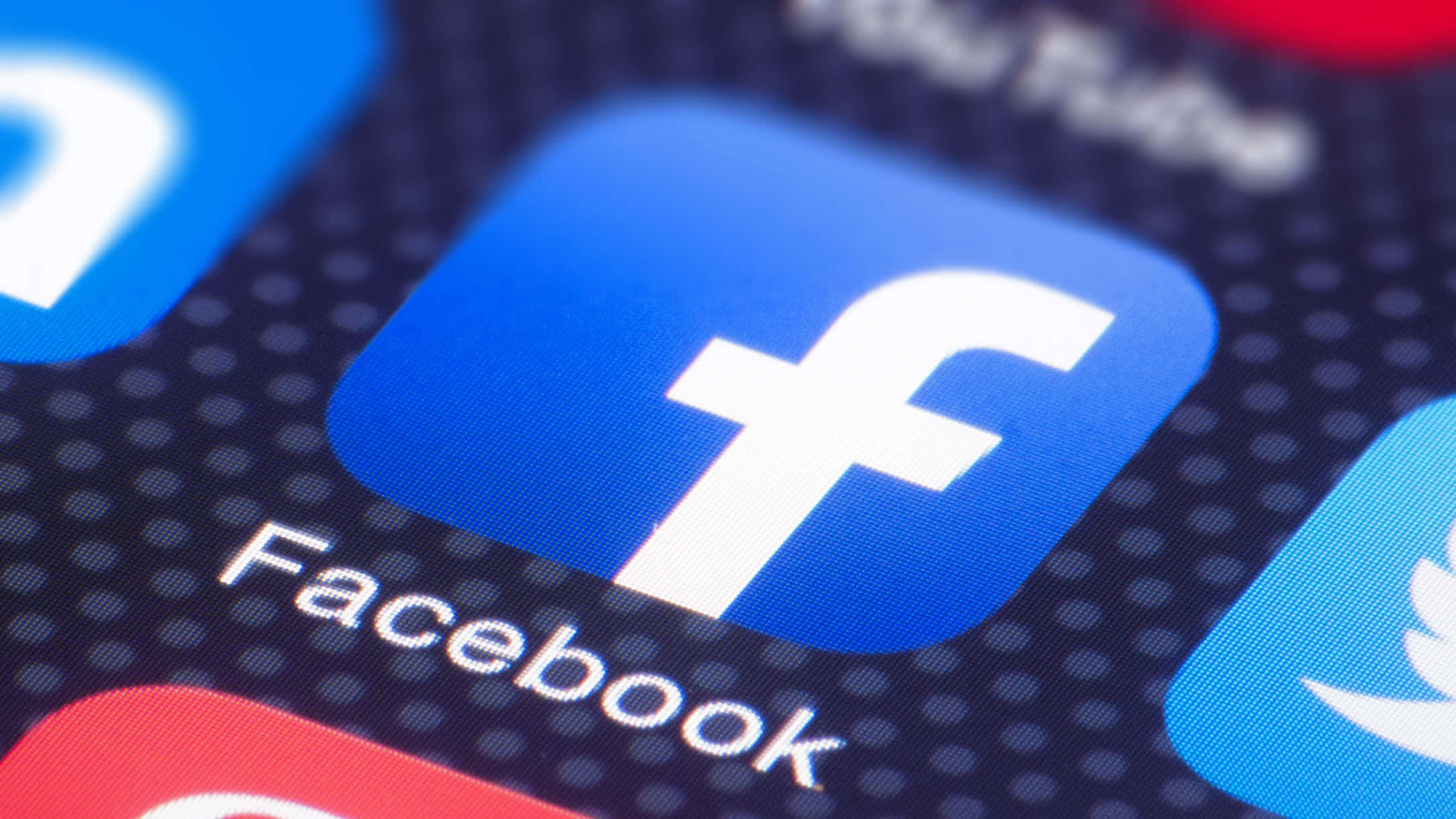 Facebook 桌布 壁紙 — 智慧型手機螢幕上藍色Facebook應用程式圖示的特寫，周圍有模糊的社交媒體圖示。