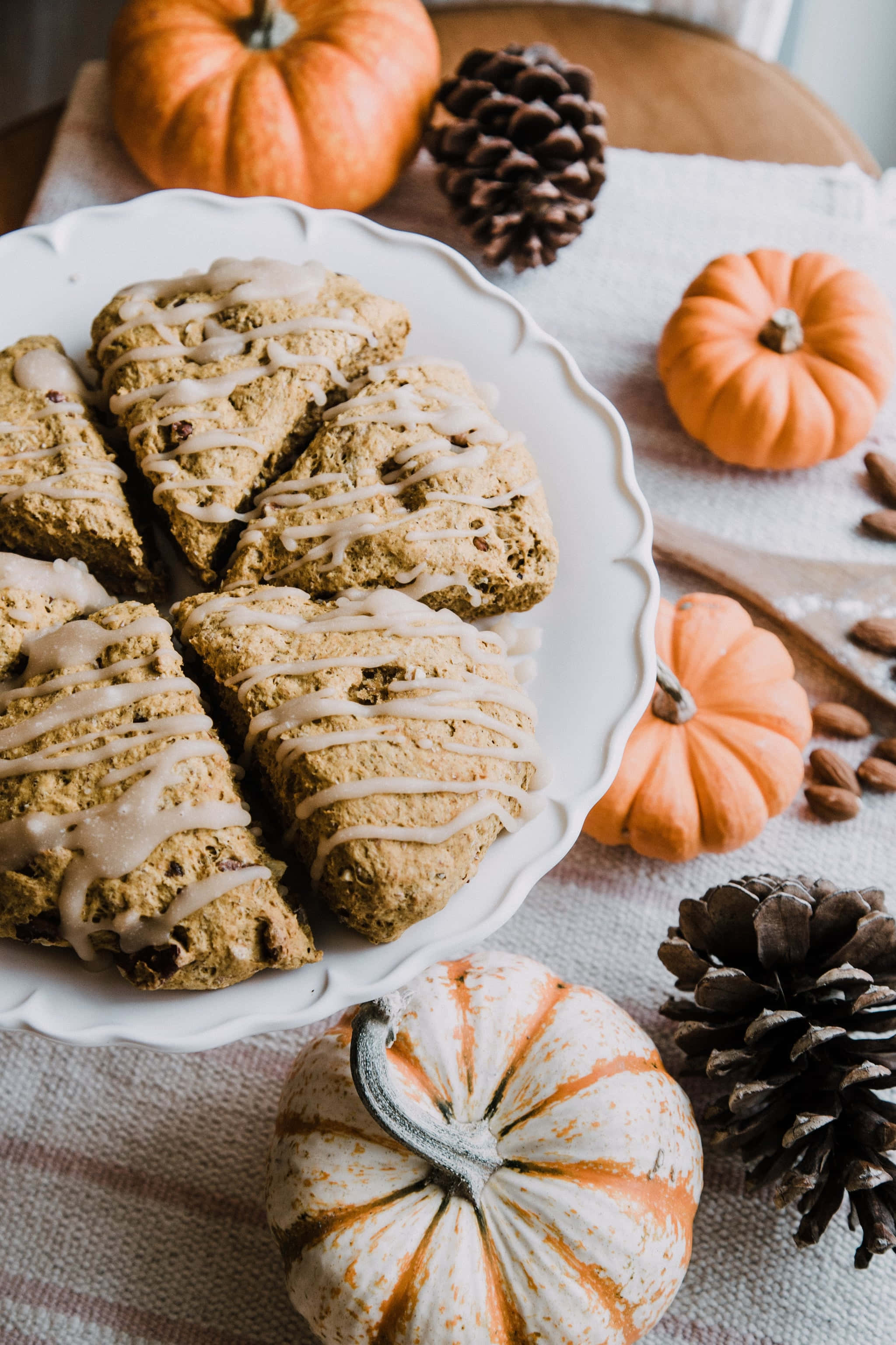 pumpkins 桌布 壁紙 — 暖南瓜司康配奶油糖霜，放在白色盤子上，周圍有小南瓜和松果，置於有紋理的布上。