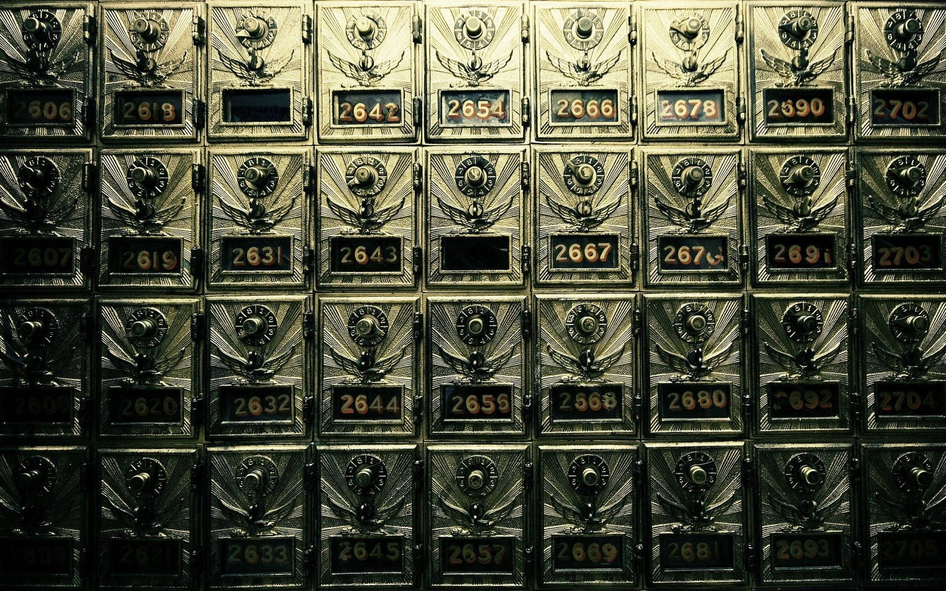 郵局 桌布 壁紙 — 暖色燈光下，復古黃銅郵箱的特寫，帶有翼形徽章和編號槽，以網格排列。