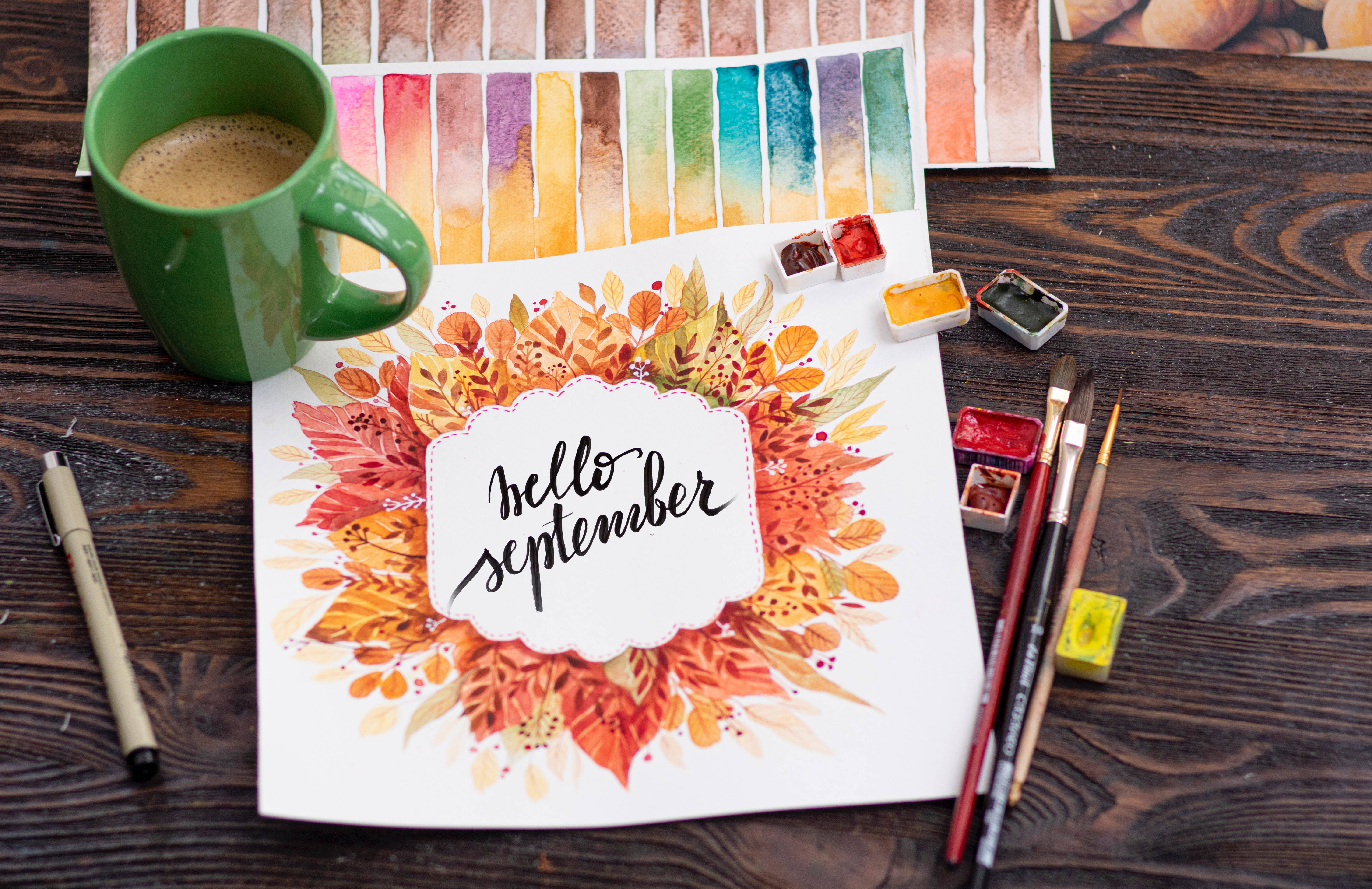 September 桌布 壁紙 — 暗色木桌上，綠色咖啡杯和美術用品旁，紙上帶有「hello september」文字的水彩秋葉花環。