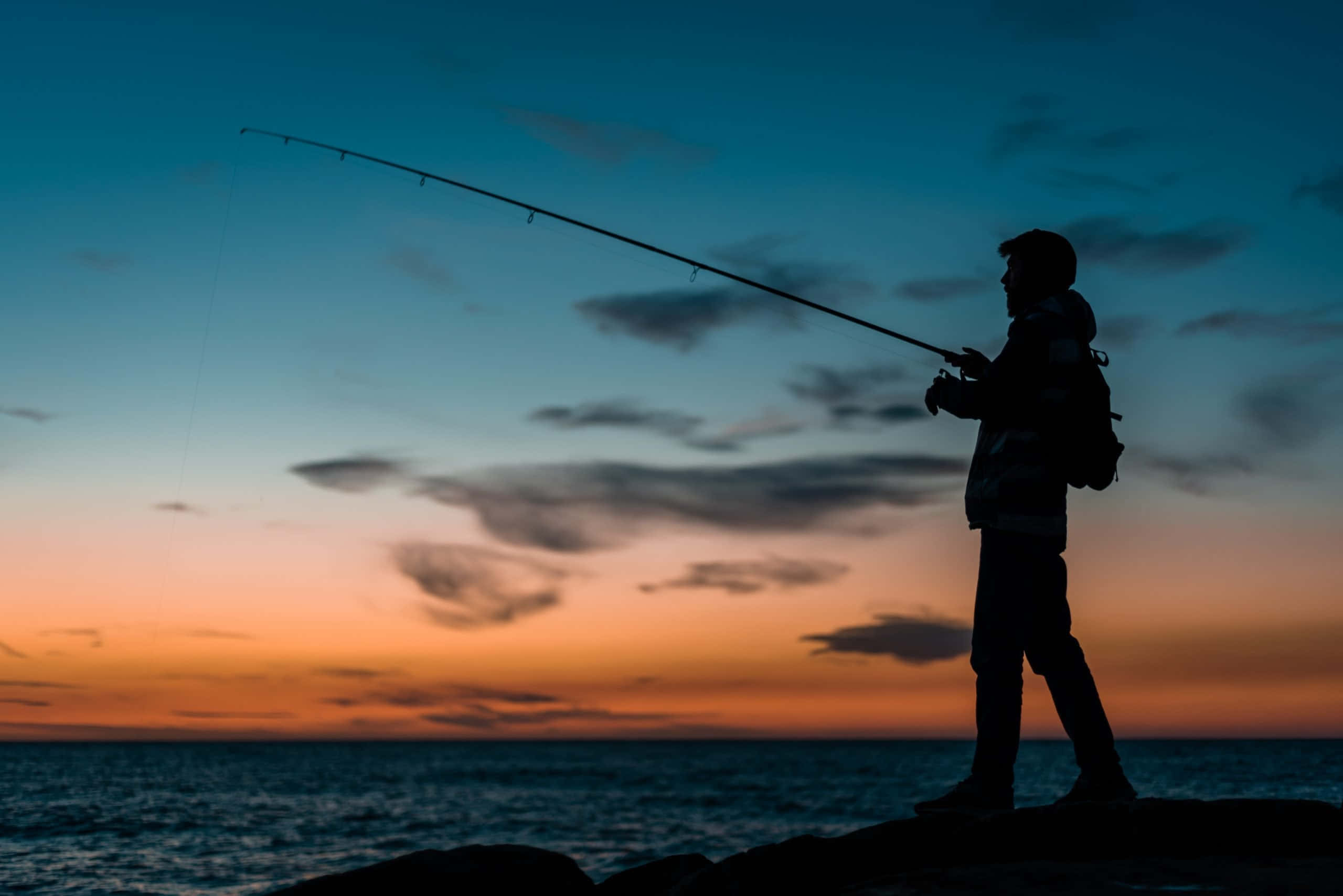 Fishing Rod 桌布 壁紙 — 暮色中，一名漁夫在岩石海岸上拋線的剪影，天空呈現鮮豔的橙色和藍色。