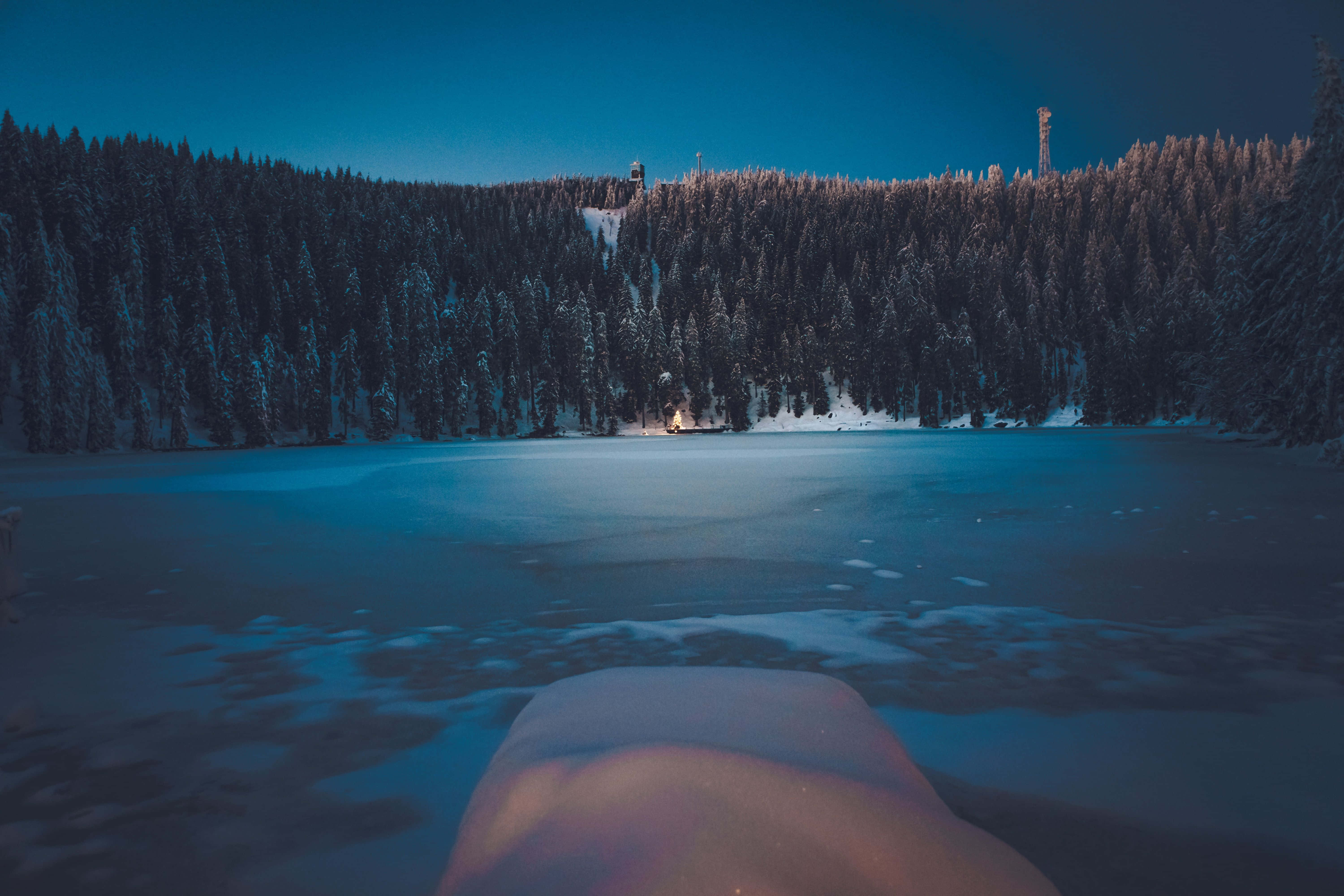 Winter Forest 桌布 壁紙 — 暮色中一個結冰的湖泊，周圍是白雪覆蓋的松樹，遠處岸邊閃爍著溫暖的光芒。
