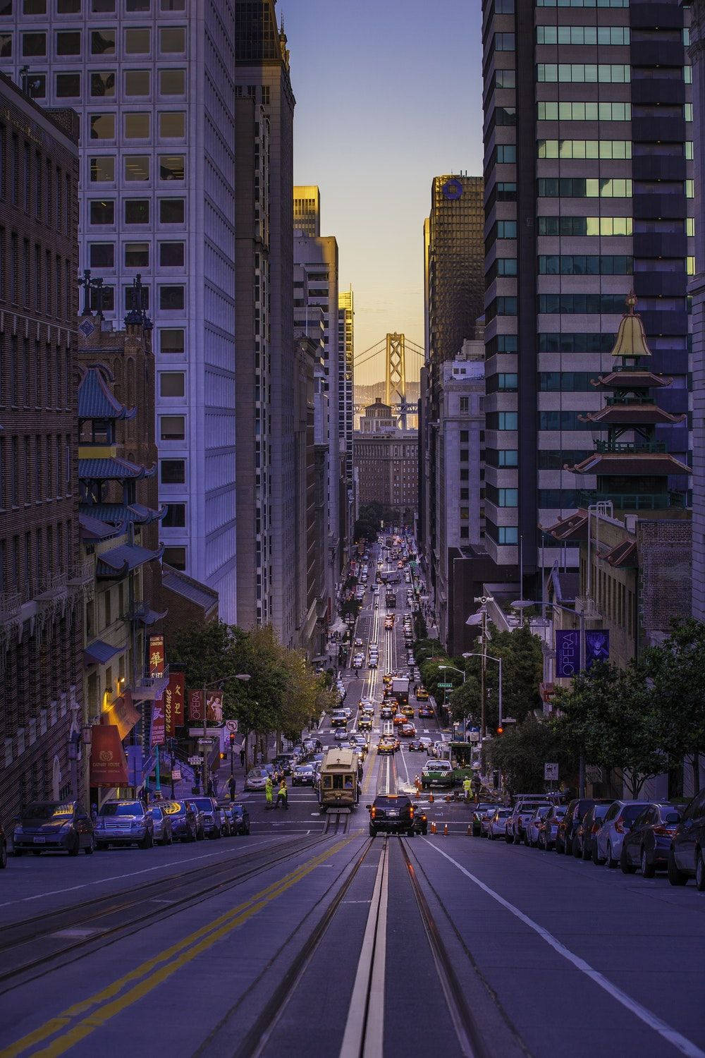 San Francisco Phone 桌布 壁紙 — 暮色中一條充滿活力的城市街道，兩旁是高聳的摩天大樓，纜車和遠處的橋樑在金色光芒中閃爍。