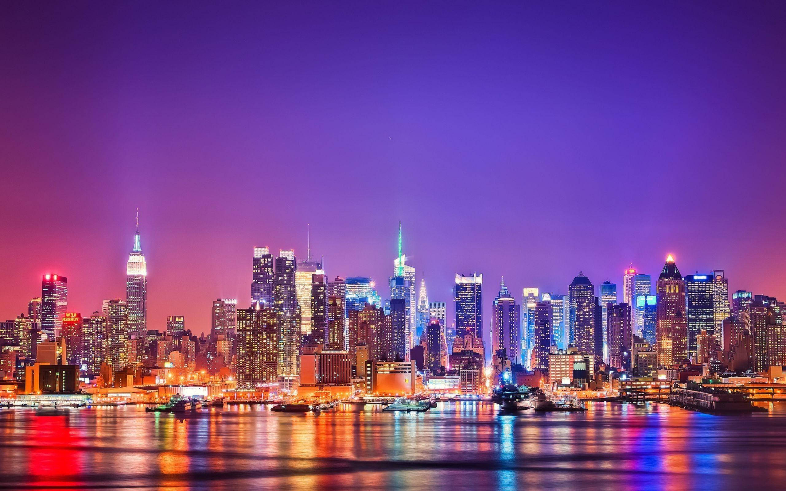 New York 桌布 壁紙 — 暮色中充滿活力的城市天際線，發光的摩天大樓倒映在平靜的水面上，天空呈現紫色。