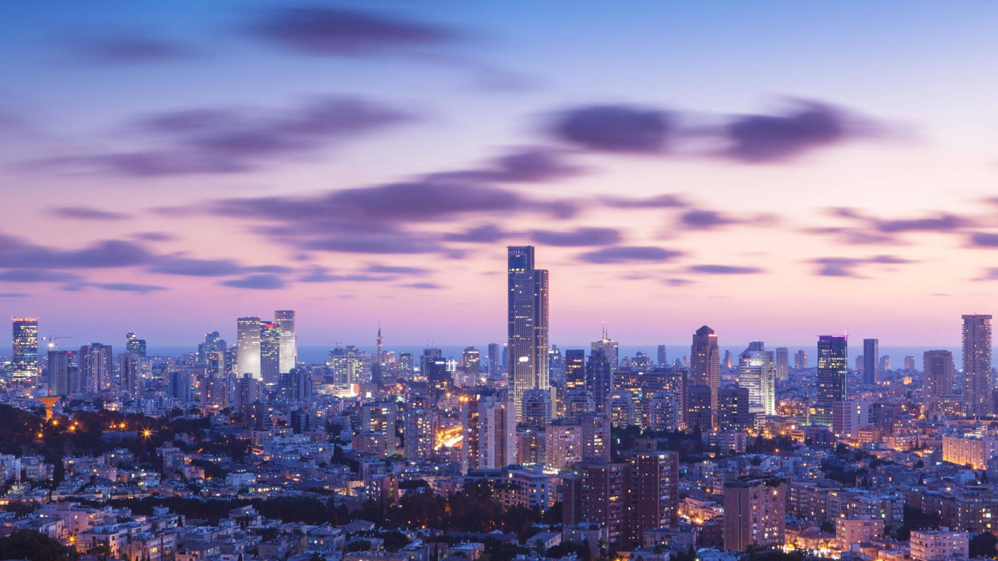 Israel 桌布 壁紙 — 暮色中廣闊的城市天際線，閃爍的摩天大樓和上方柔和的紫粉色雲彩。