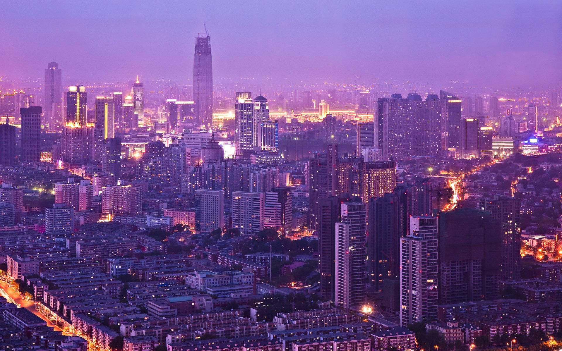 Tianjin 桌布 壁紙 — 暮色中廣闊的城市景觀，摩天大樓在紫色天空下閃耀，街道被溫暖的金色燈光照亮。