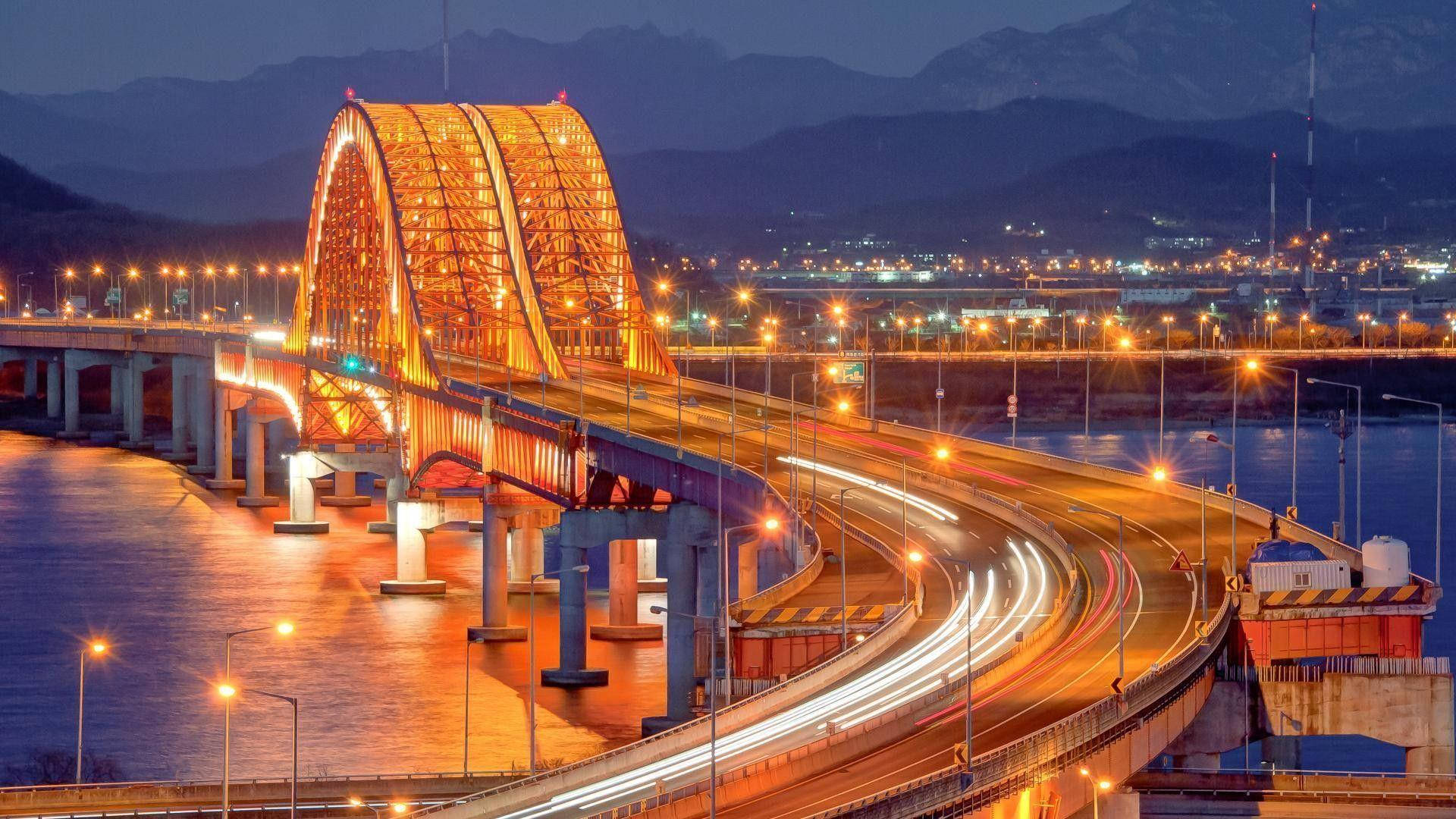 南韓 桌布 壁紙 — 暮色中發光的拱橋，水面上有汽車燈光軌跡，遠山依稀可見。