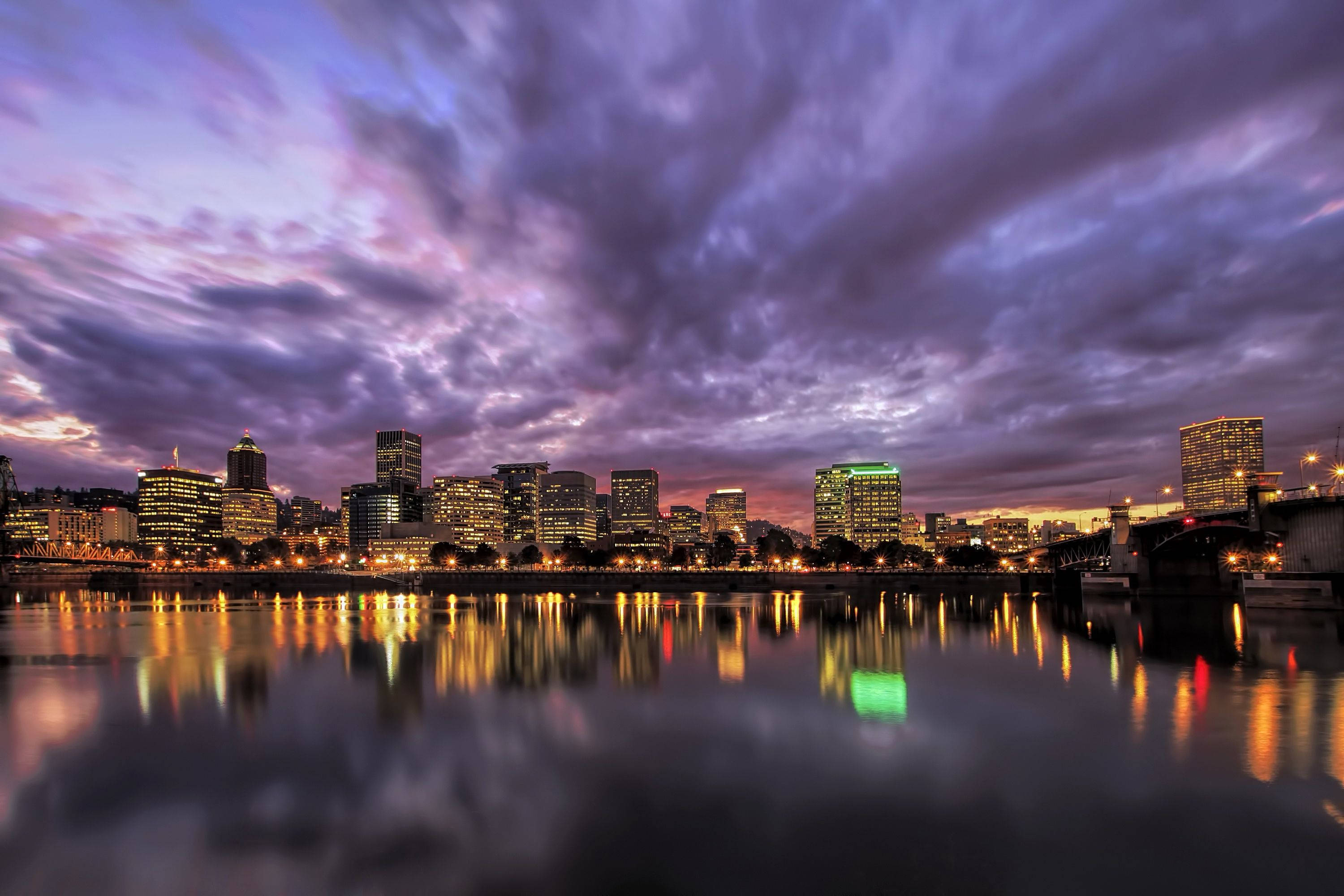 Portland 桌布 壁紙 — 暮色中的城市天際線，帶有紫色雲彩和寧靜水面上閃爍的建築倒影。