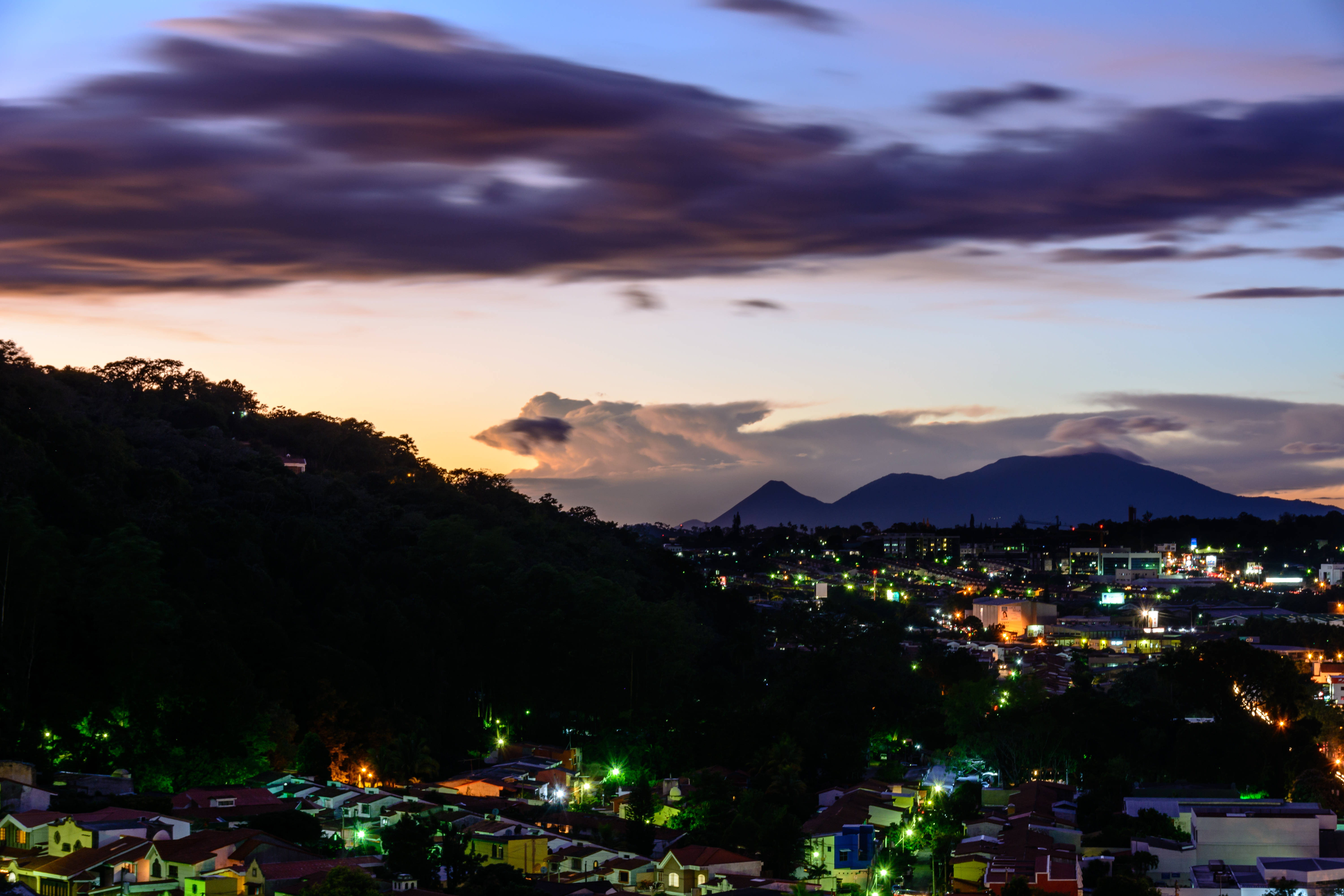 El Salvador 桌布 壁紙 — 暮色中的城市景觀，燈光閃爍，坐落在黑暗的山丘和遠處的山脈之間，天空呈現紫色和橙色。