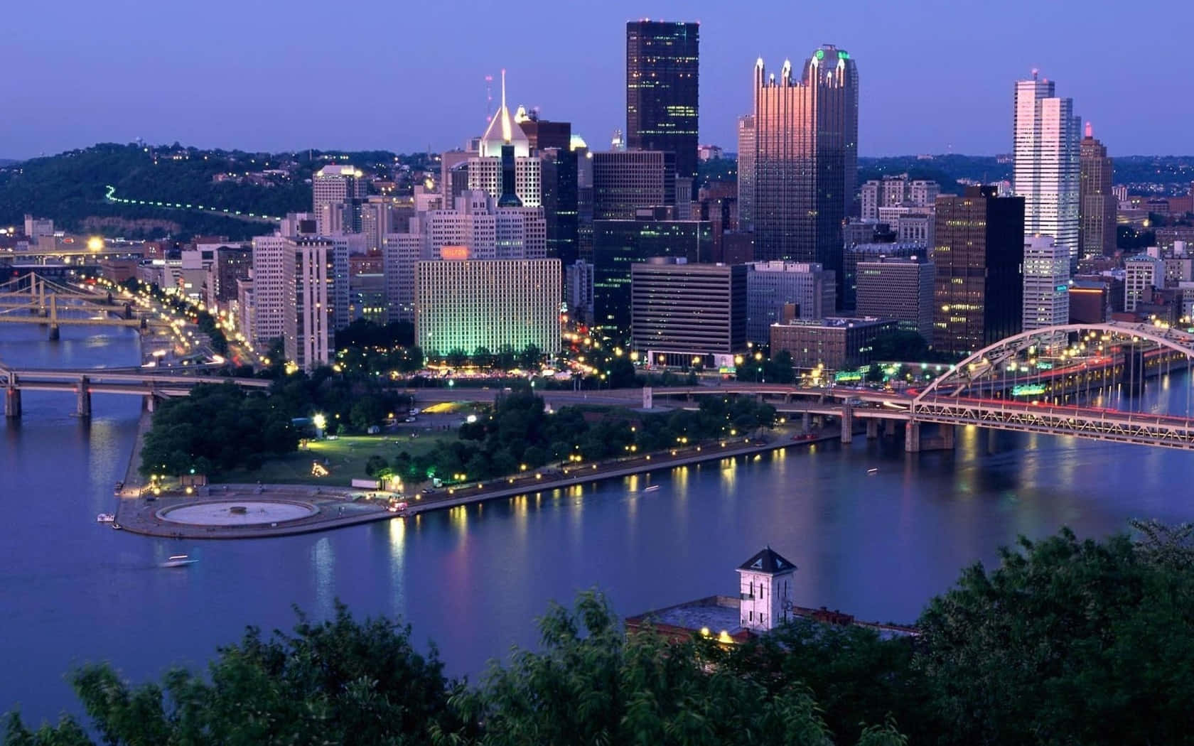 Pittsburgh Skyline 桌布 壁紙 — 暮色中的城市景觀，燈火通明的摩天大樓和橋樑倒映在平靜的河面上，四周環繞著綠意。