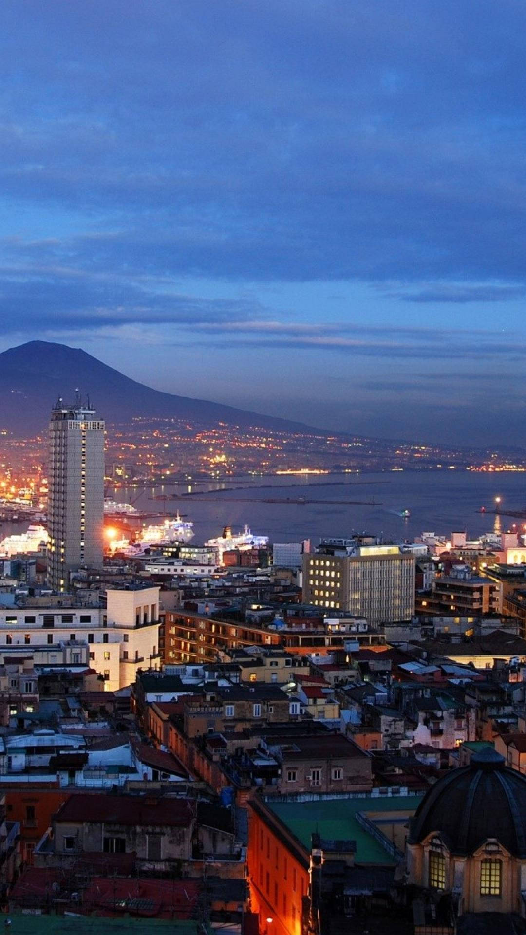 Naples 桌布 壁紙 — 暮色中的城市景觀，發光的建築物和一座黑暗的山脈籠罩著港口。