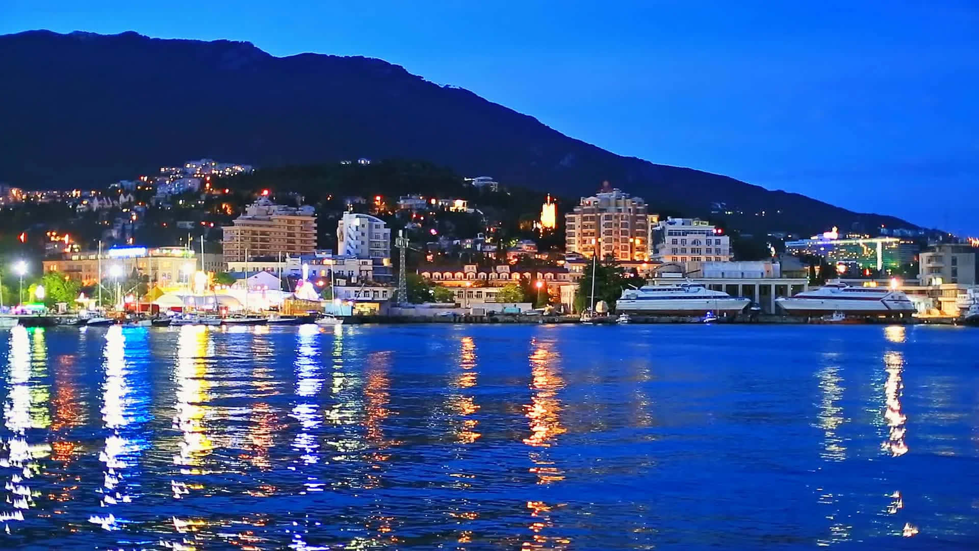 City Lights 桌布 壁紙 — 暮色中的沿海城市，燈光閃爍，倒映在平靜的水面上，以黑暗的山脈為背景。